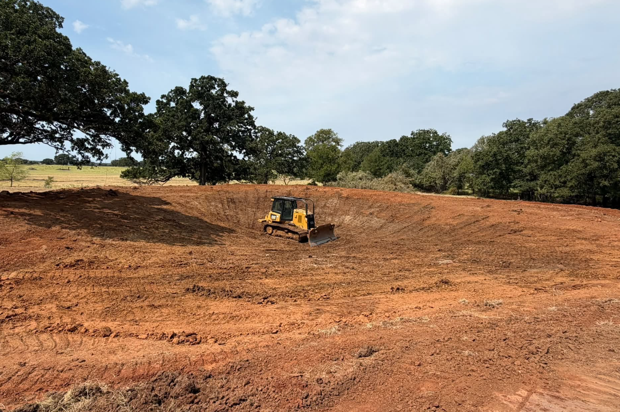 A construction site with a yellow bulldozer on red soil, surrounded by green trees and a blue sky with some clouds.