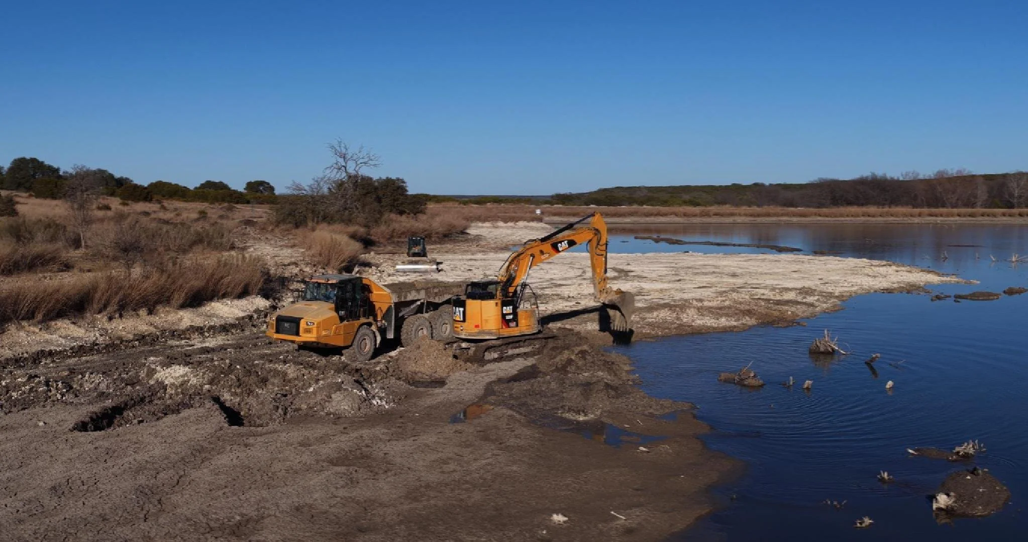 Construction machinery operating along the shoreline of a body of water, with clear blue sky and sparse trees and bushes in the background.