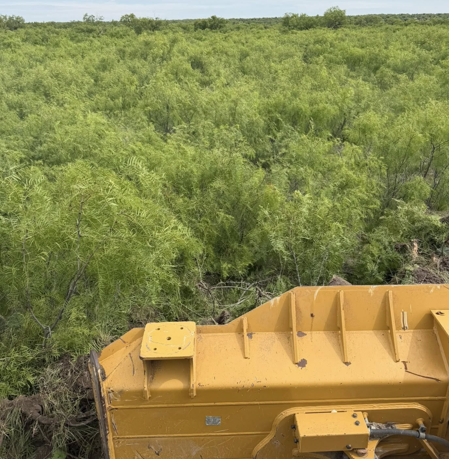 A yellow construction or agricultural machine part situated near a lush green forest, under a cloudy sky.