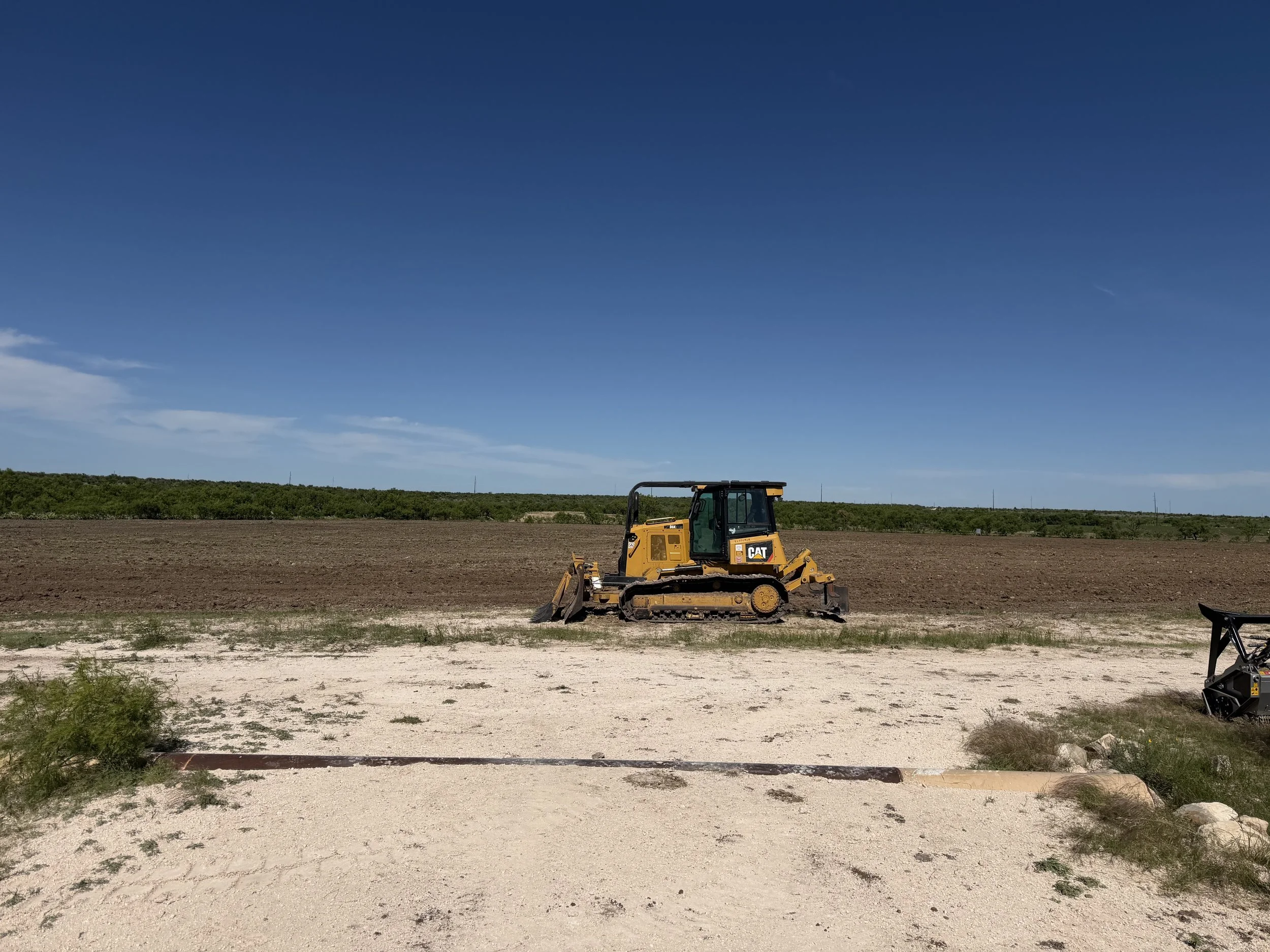 A yellow Caterpillar bulldozer working on a dirt field under a clear blue sky. In the background, there is green shrubbery and a flat horizon.