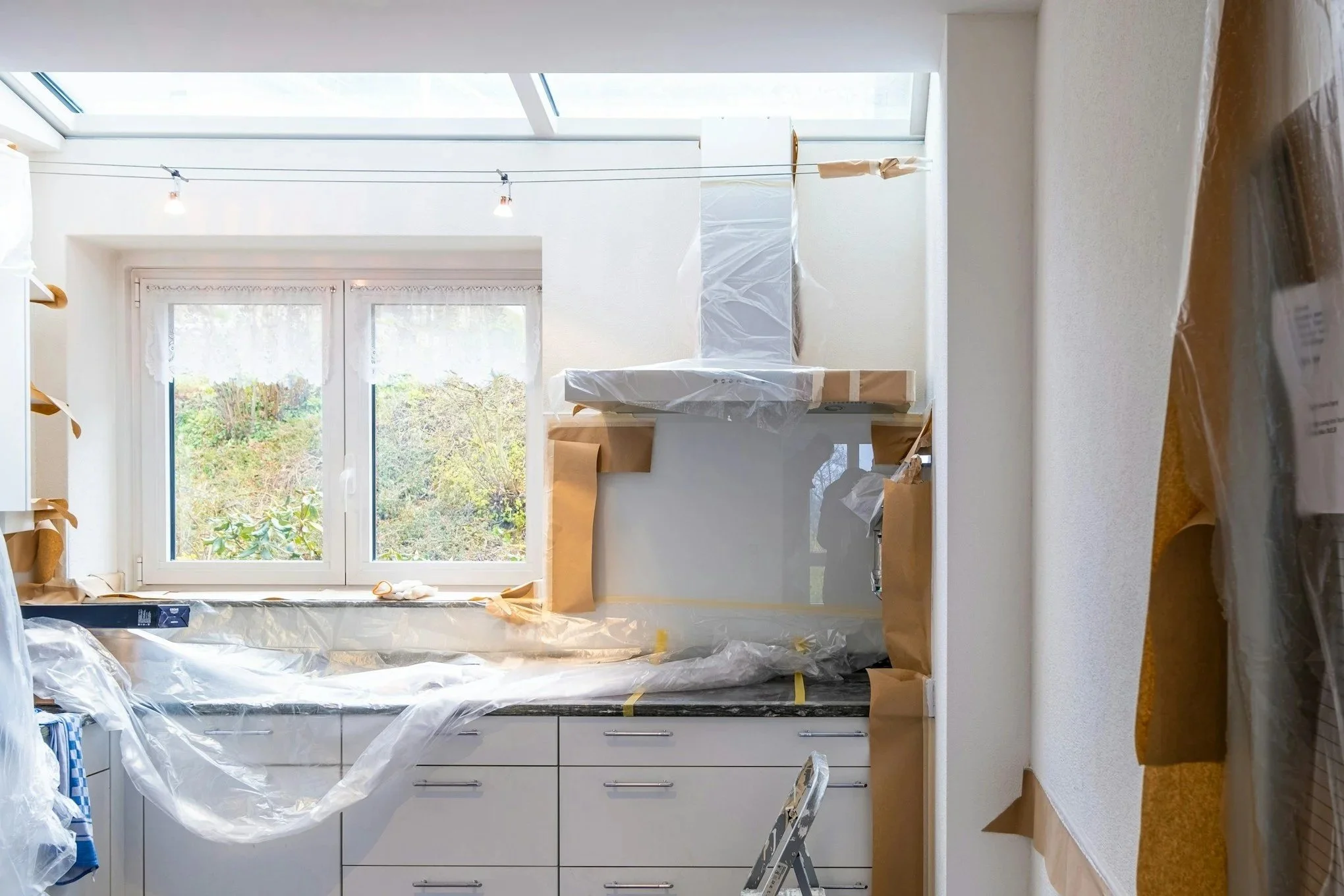 A kitchen undergoing renovation, with cabinets covered in plastic, a ladder, and construction materials scattered around. It has large windows and a skylight letting in natural light.