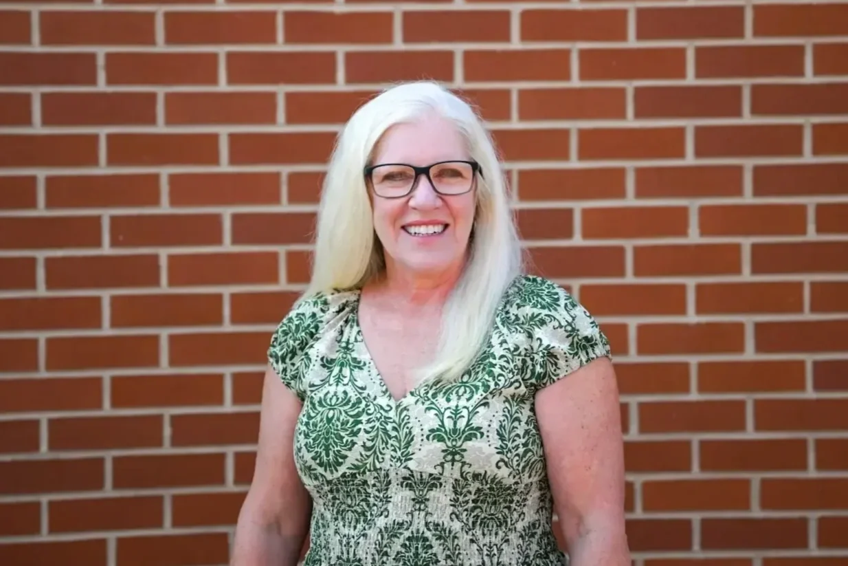 A smiling woman with long white hair, wearing glasses and a green patterned dress, standing in front of a brick wall.