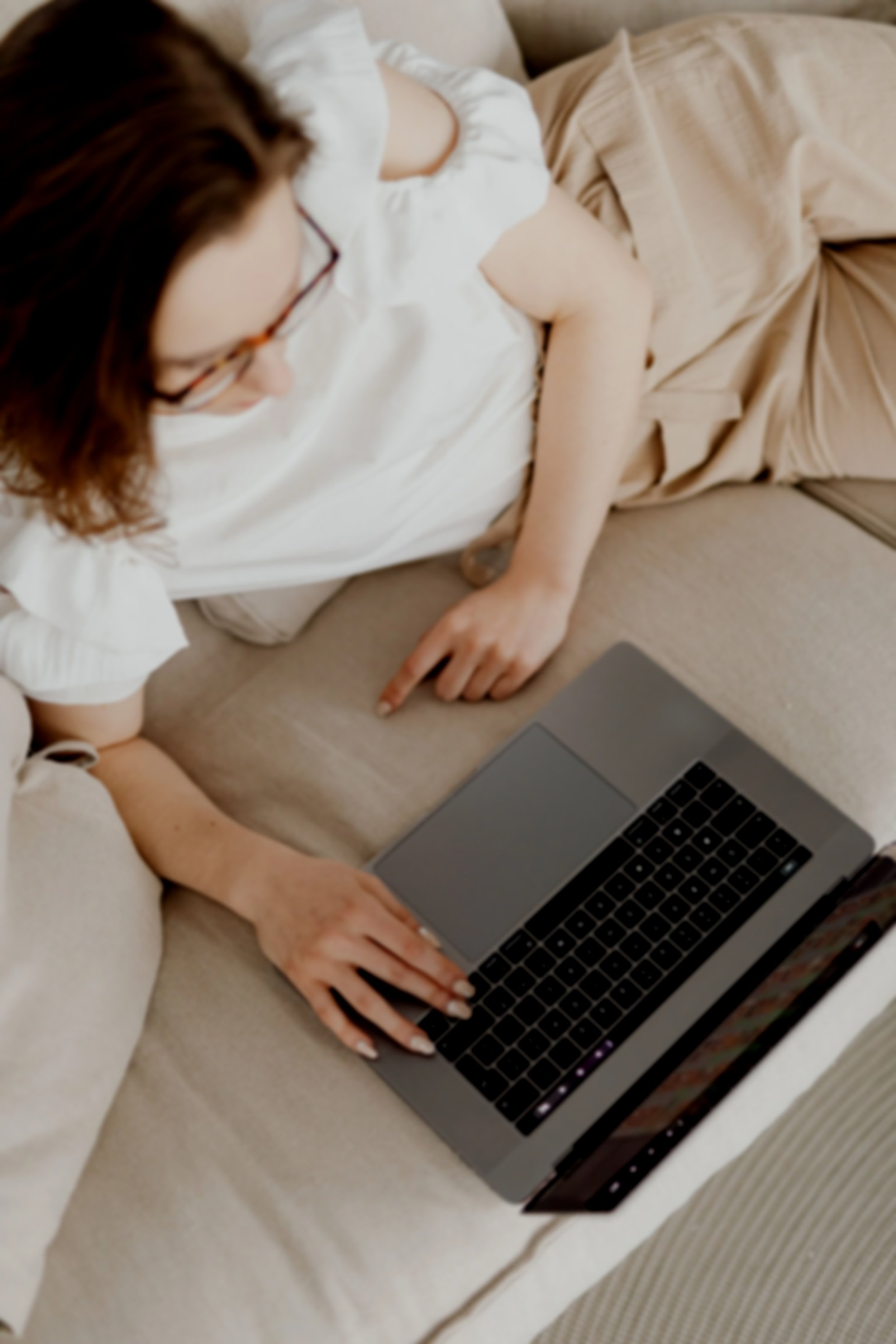 A person with glasses working on a laptop while lying on a beige sofa, with a blanket and pillows around.