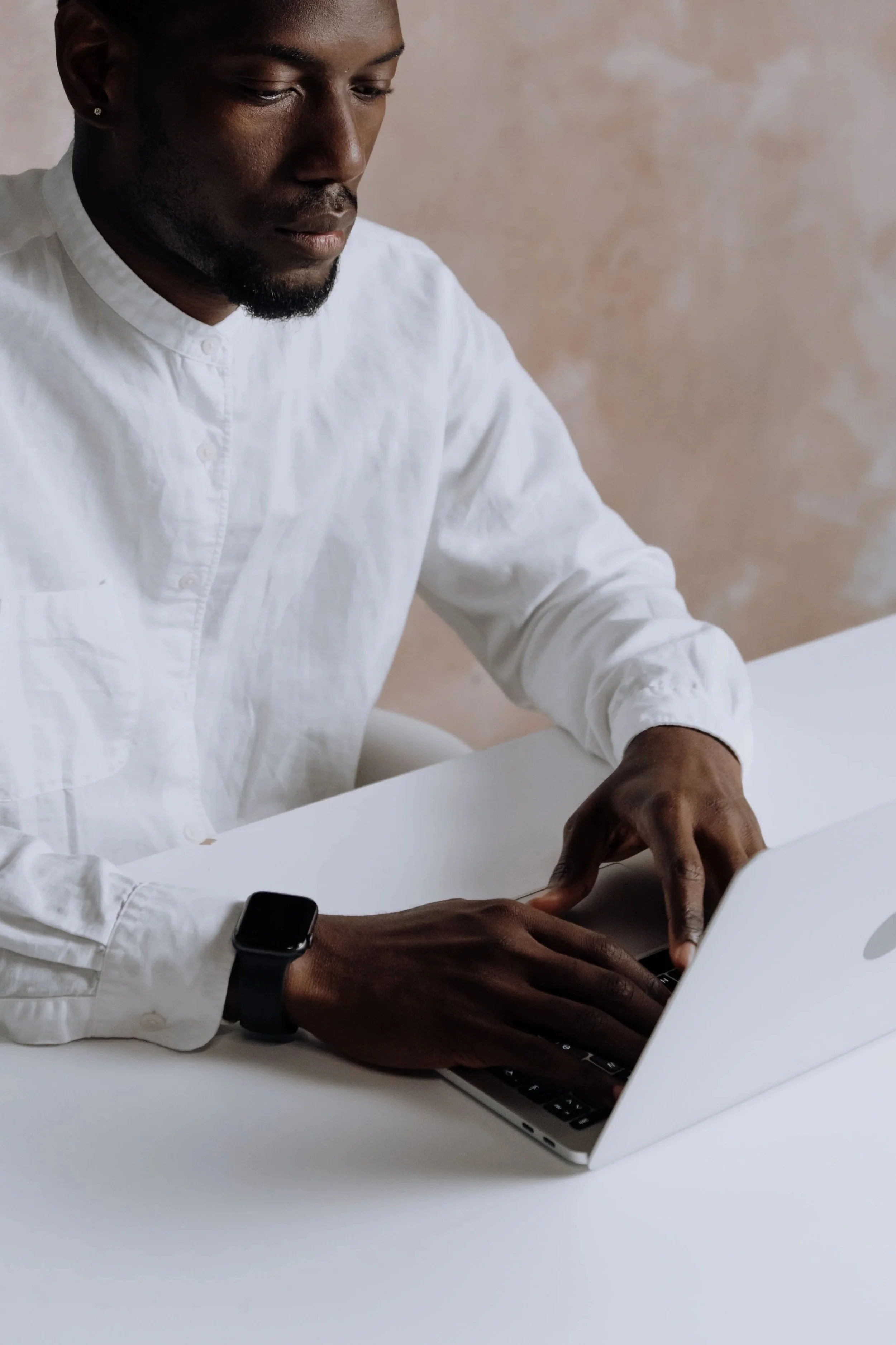 A person with dark skin, wearing a white shirt and a smartwatch, working on a laptop at a white desk