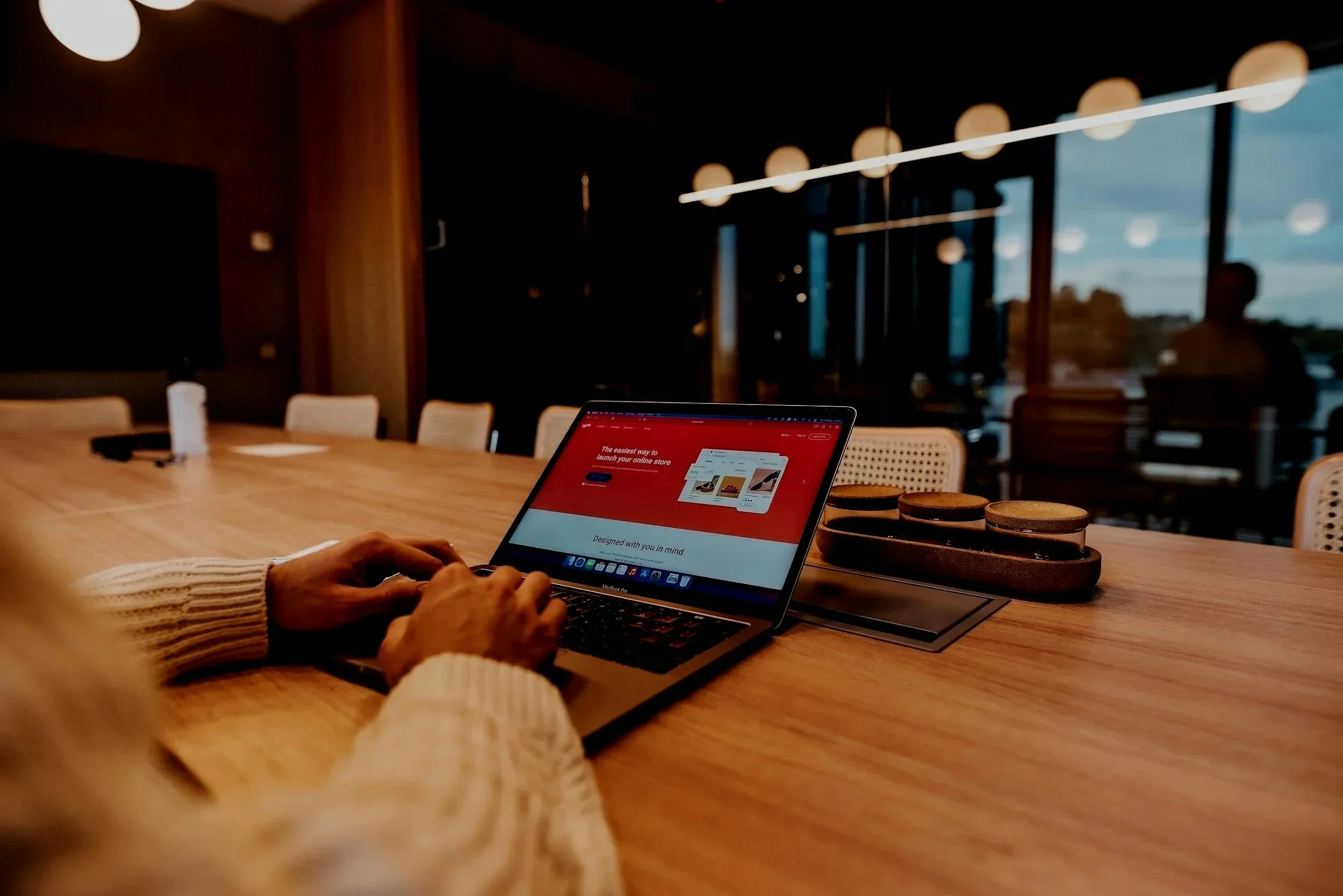 Person using a laptop on a wooden table in a modern meeting room with large windows and hanging lights.