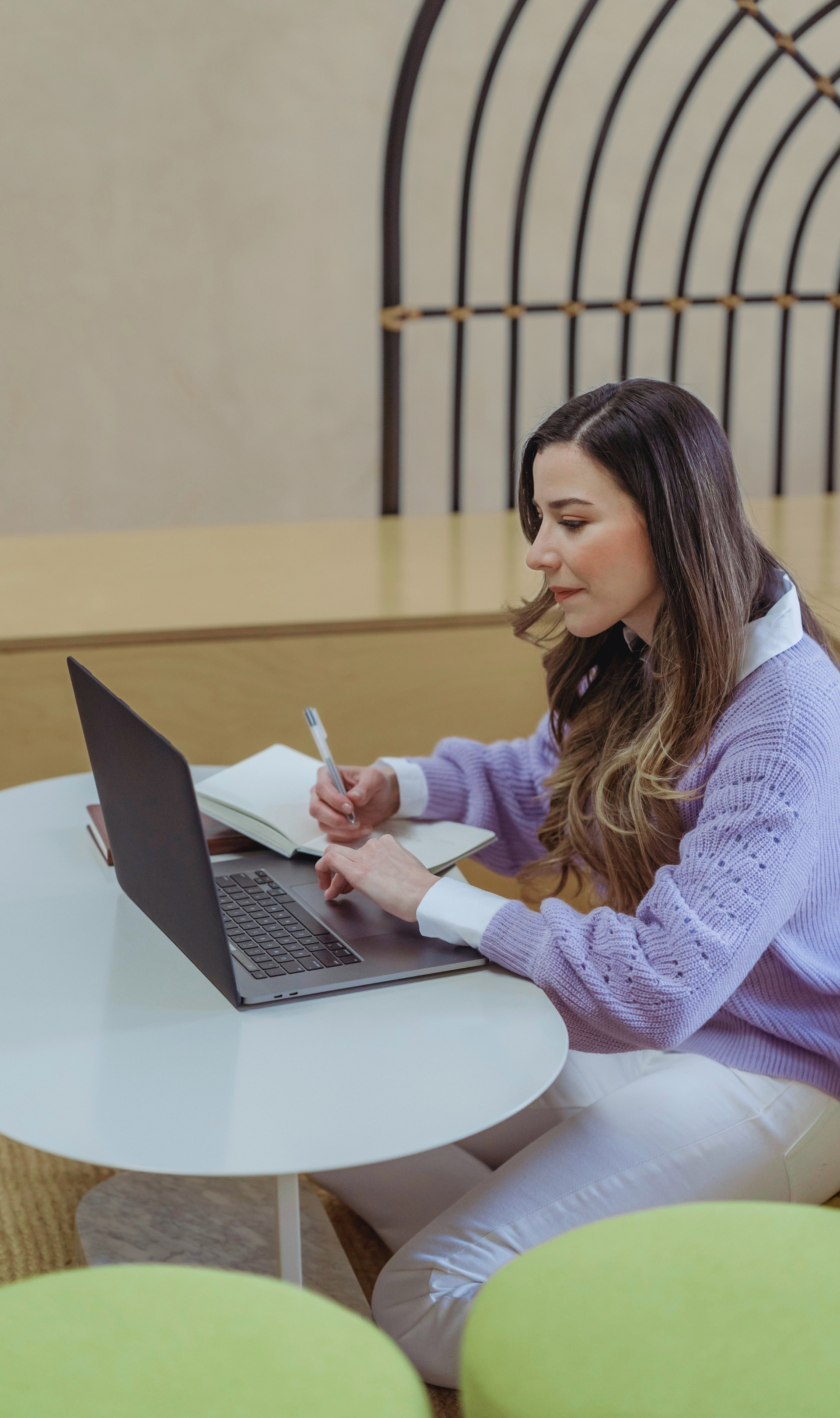 A woman with long brown hair sitting at a white round table, working on a laptop, with a notebook and pen in her hands, in a modern room with green chairs in the foreground and a decorative wall feature in the background.