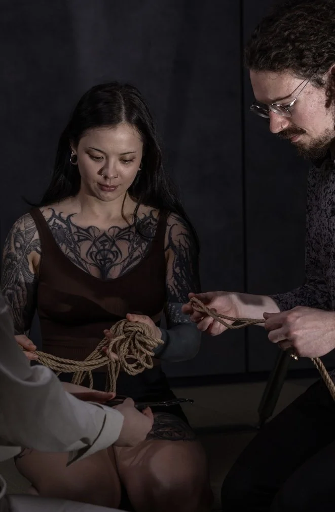 A woman with tattoos being tied up with rope by two men in a dark room.