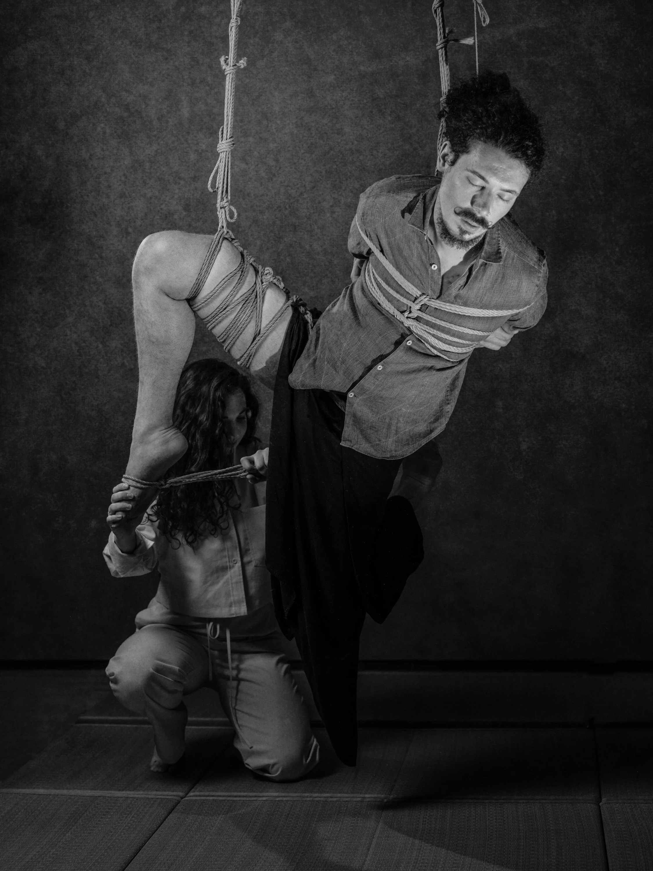 A person is bound in ropes, with another person kneeling and holding one of the person's wrists, against a plain dark background.