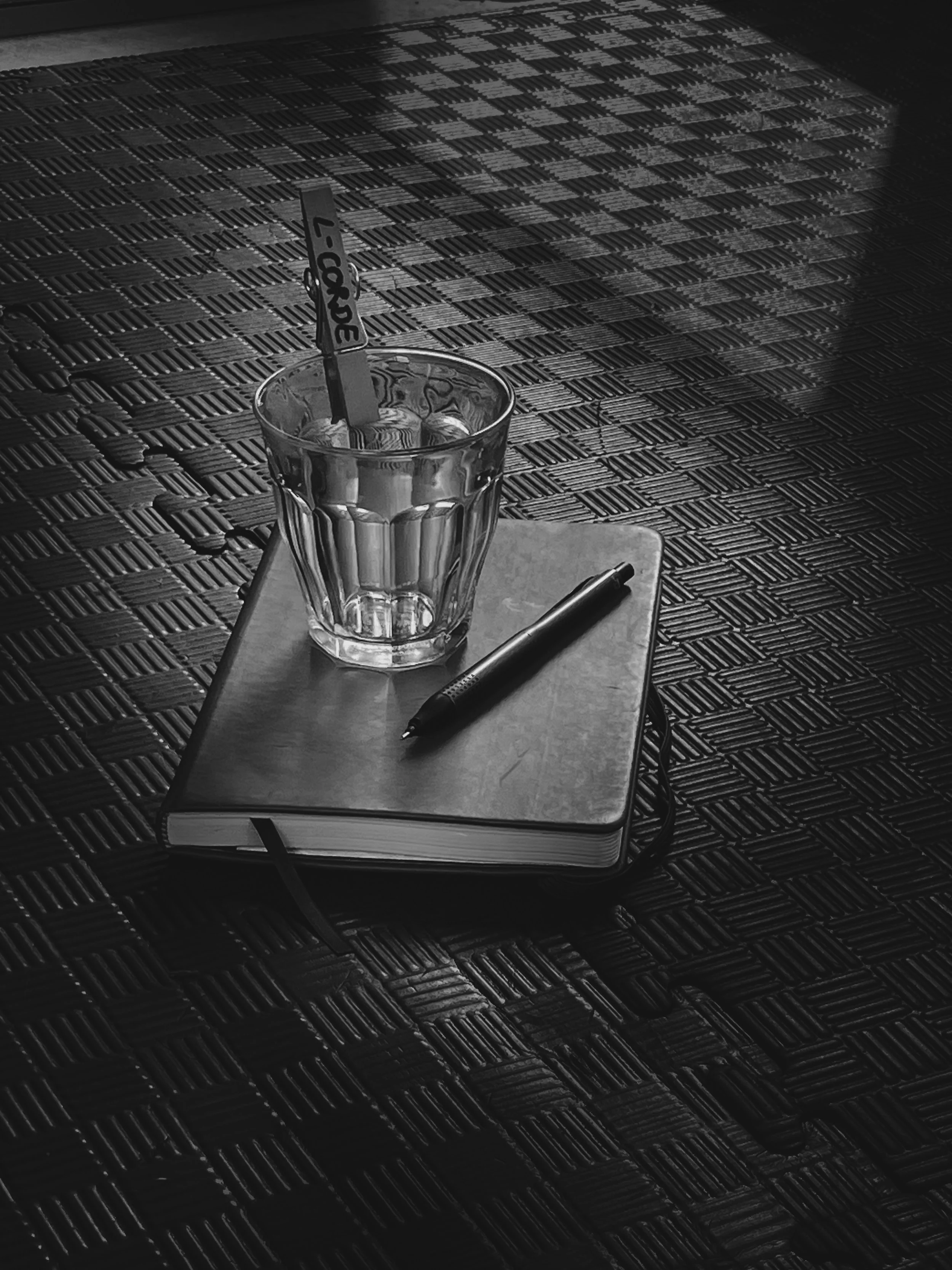 A glass of iced water with a clip, a notebook with a pen on top, on the floor, with a textured floor in the background.
