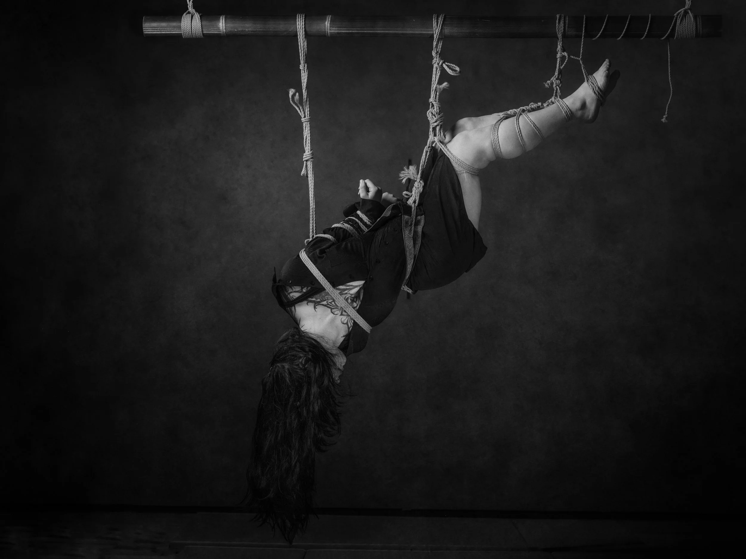 A person with long hair is hanging upside-down from a horizontal wooden bar, wrapped in ropes for a bondage pose, with a dark background.