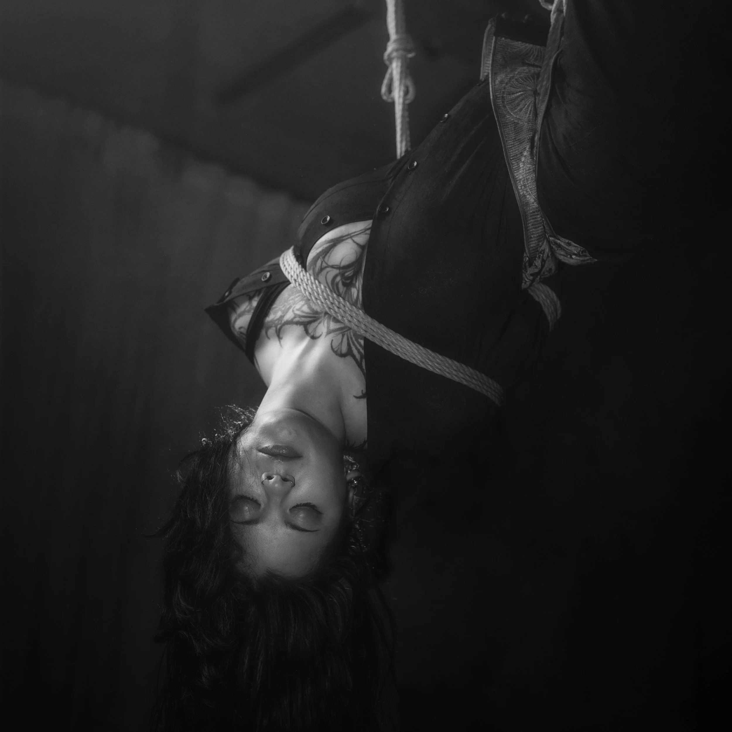 A woman lying down with her eyes closed, with long dark hair, nose piercing, and tattoos on her arm, wearing a sleeveless top, in a dark setting.