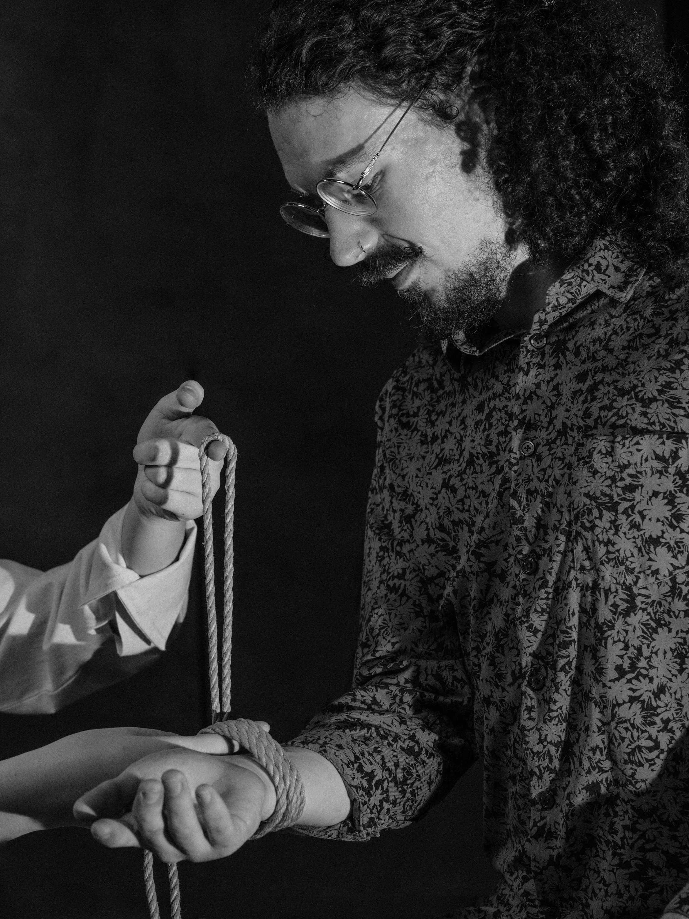 A man with curly hair, glasses, and a floral shirt is looking down at his arm, which is being held by another person. The person's hand is wrapped with a rope, and the scene appears to be in black and white.