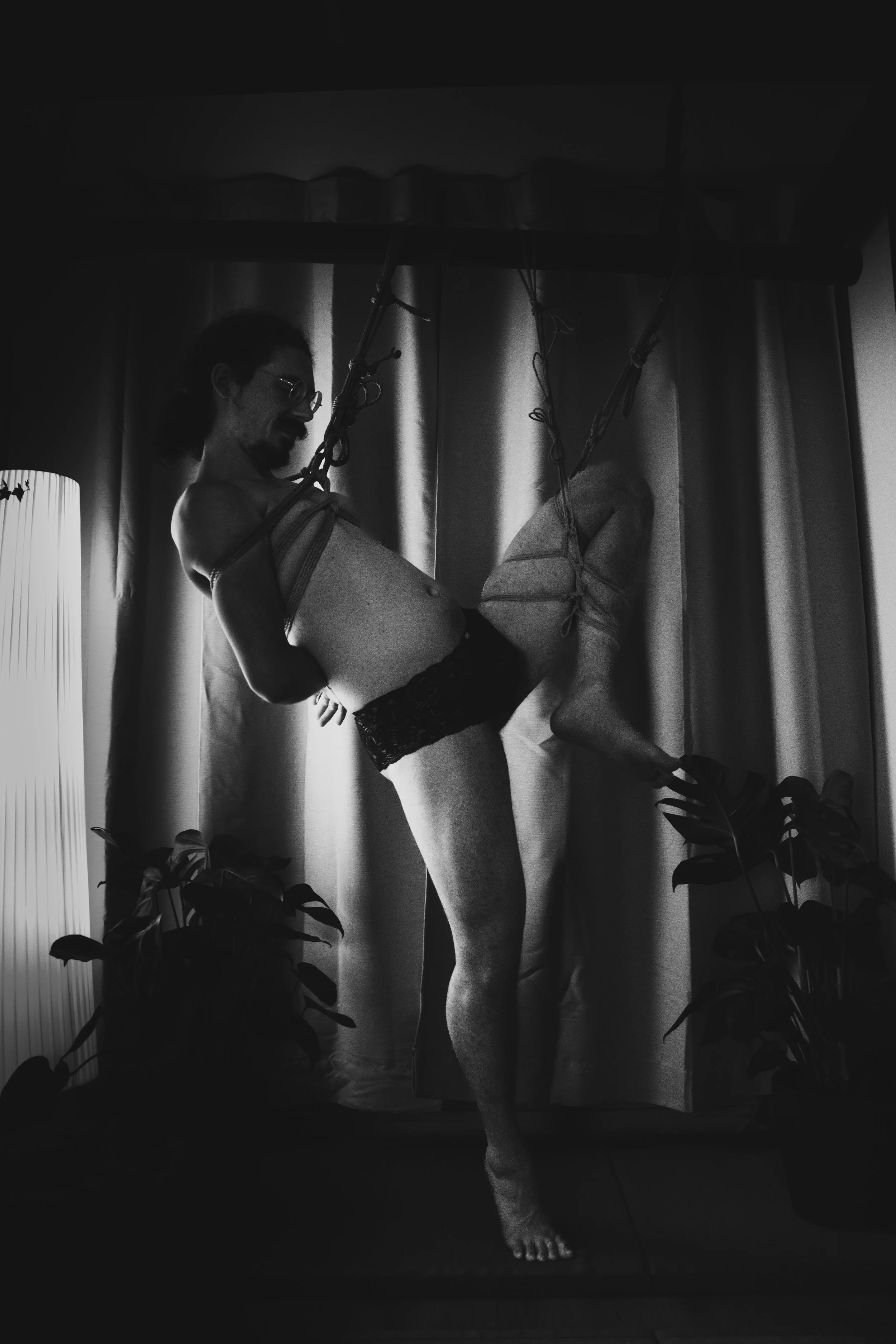 A person with long hair, glasses, and a beard is in a shibari partial suspension