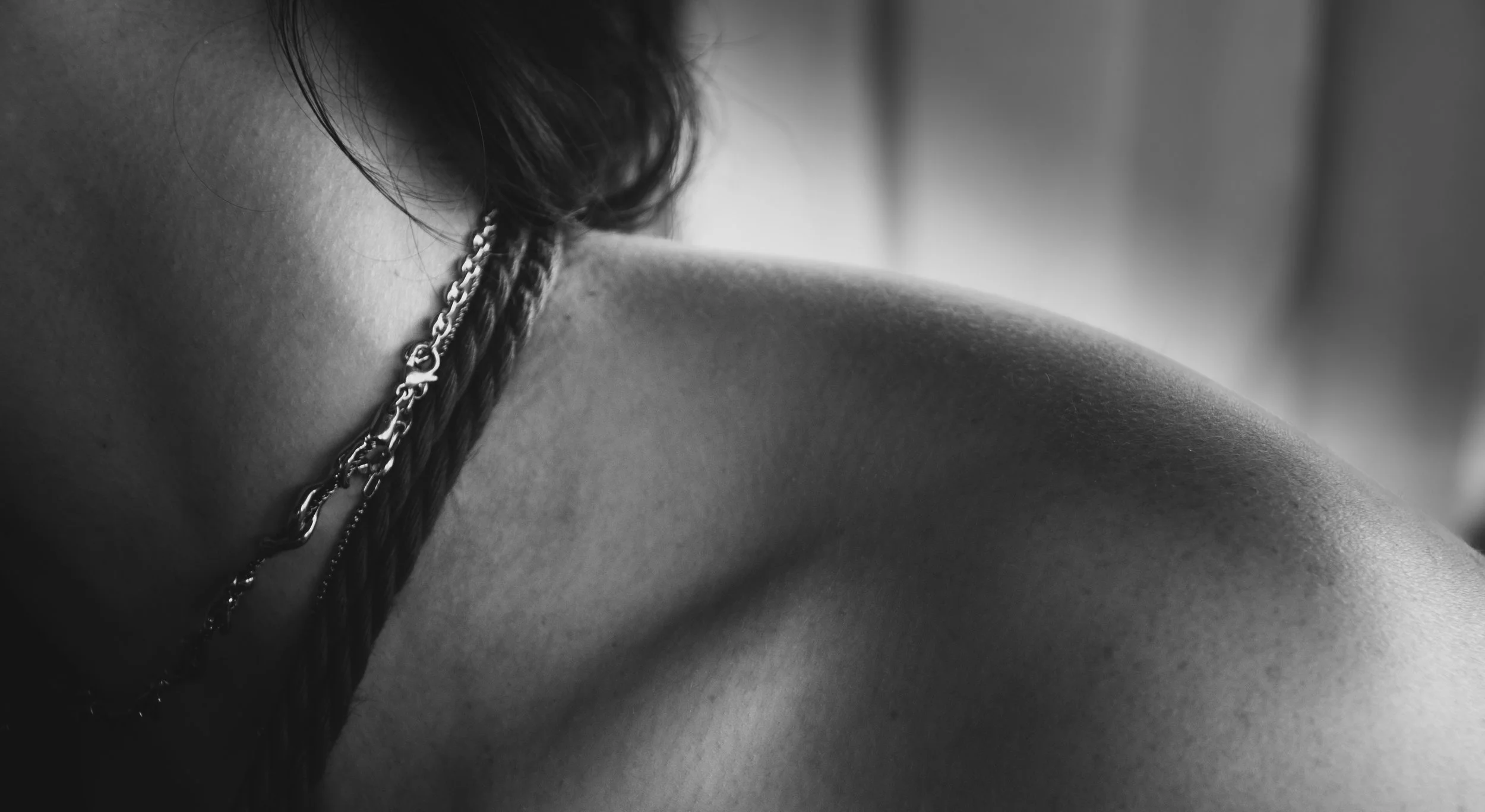 Close-up black and white photograph of a person's shoulder with a chain necklace, with the person's hair partially covering the neck.