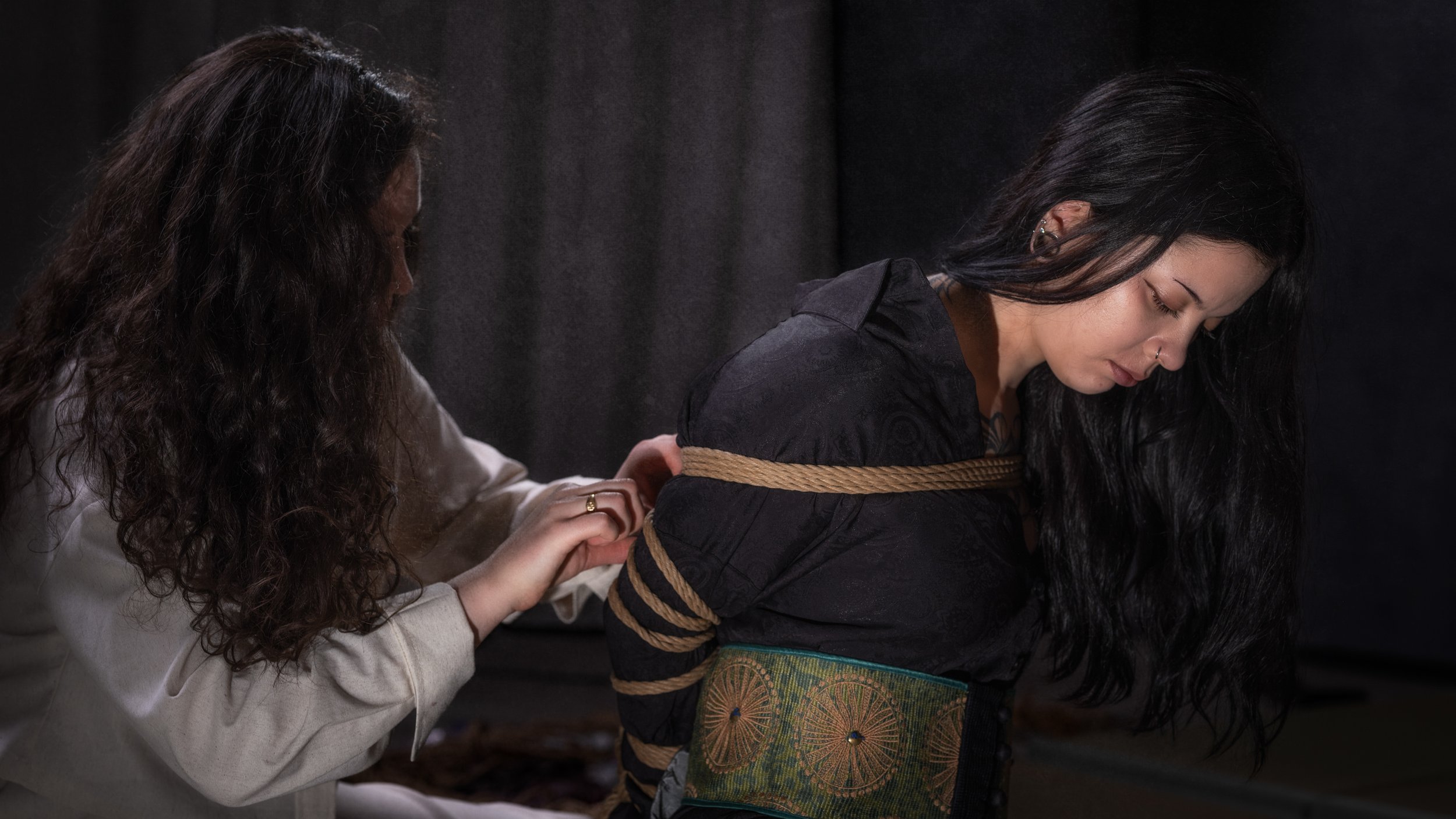 A woman with black hair and a nose piercing is tied up with rope and looking down, while another woman with long, curly hair is holding her arm and appears to be helping her.