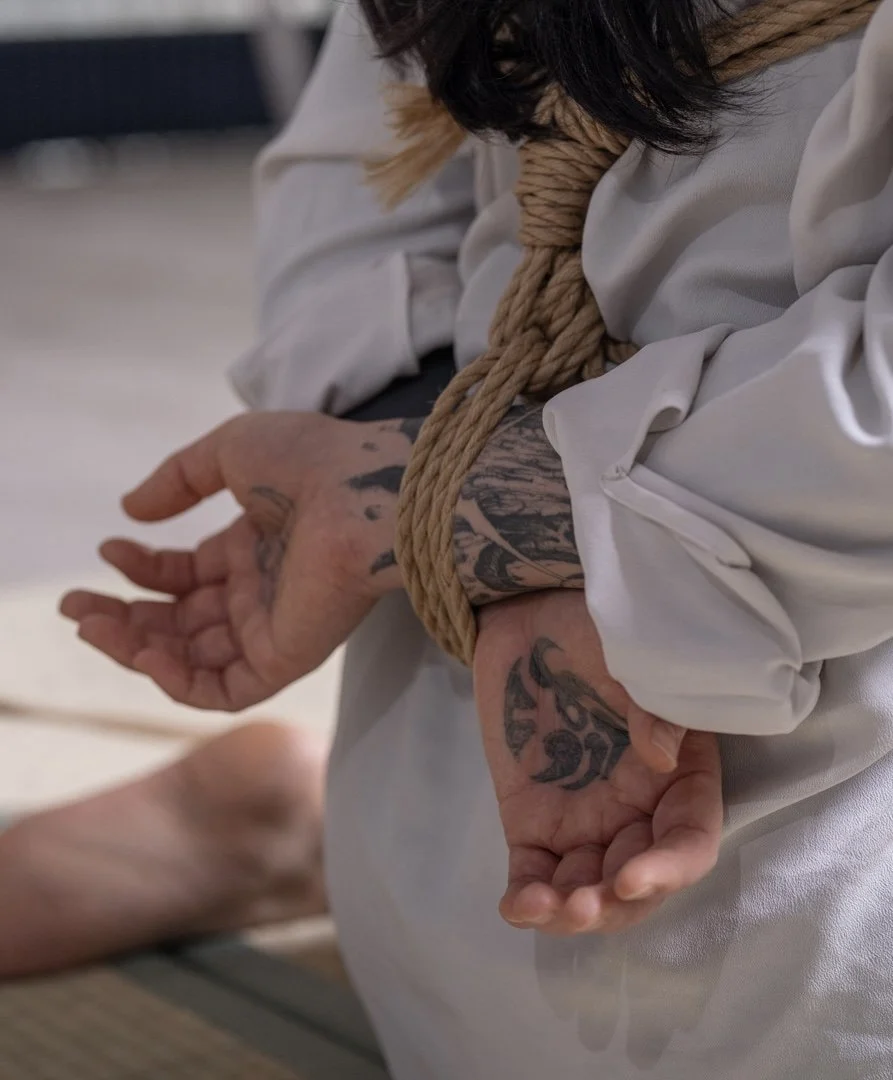 Person with tattoos on their hand and arm, dressed in a white outfit, tied with a rope around the torso, kneeling on the floor.