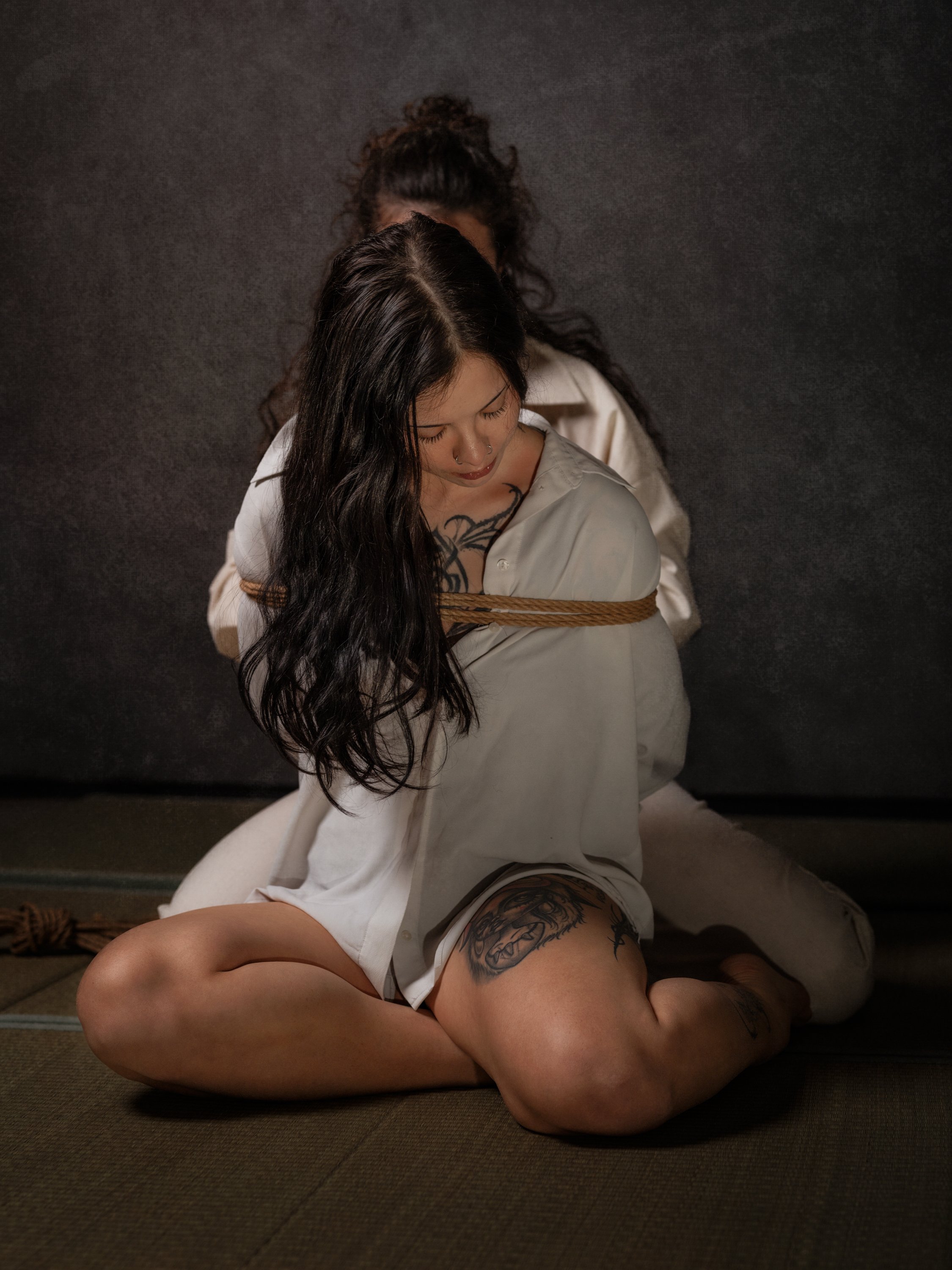 Two women with tattoos are tied up with rope and sitting on the floor against a black wall.