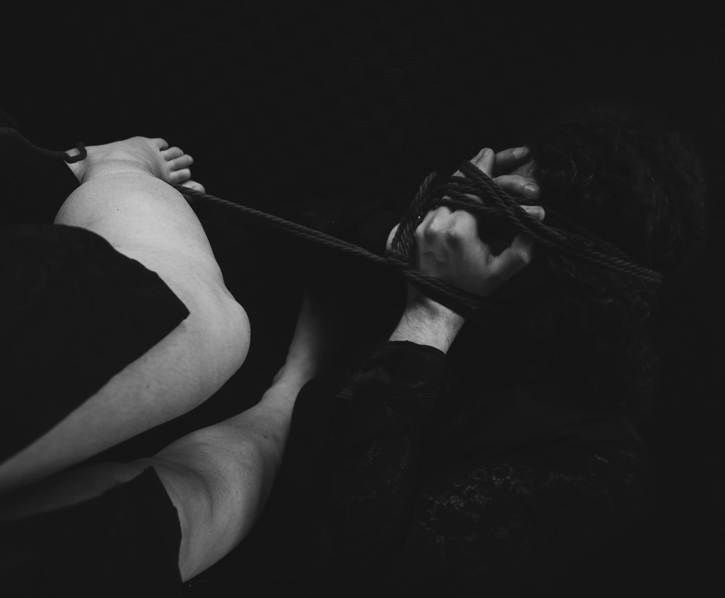 A black and white photo of a person lying in on the floor while being tied. legs of the rigger visible, with dark textured flooring beneath.