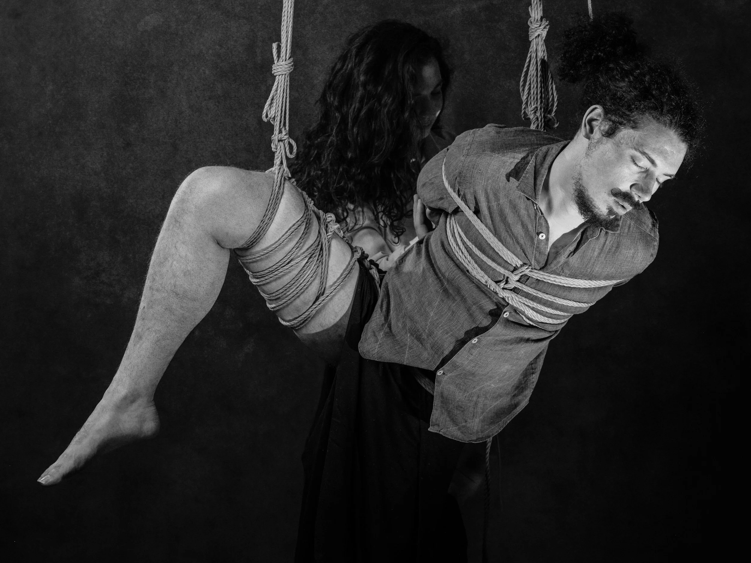 A black and white photo of a man and woman, with the woman behind the man, both bound and leaning forward. The woman appears to be holding the man's arm while they are suspended with ropes in what seems to be a staged or artistic setting.