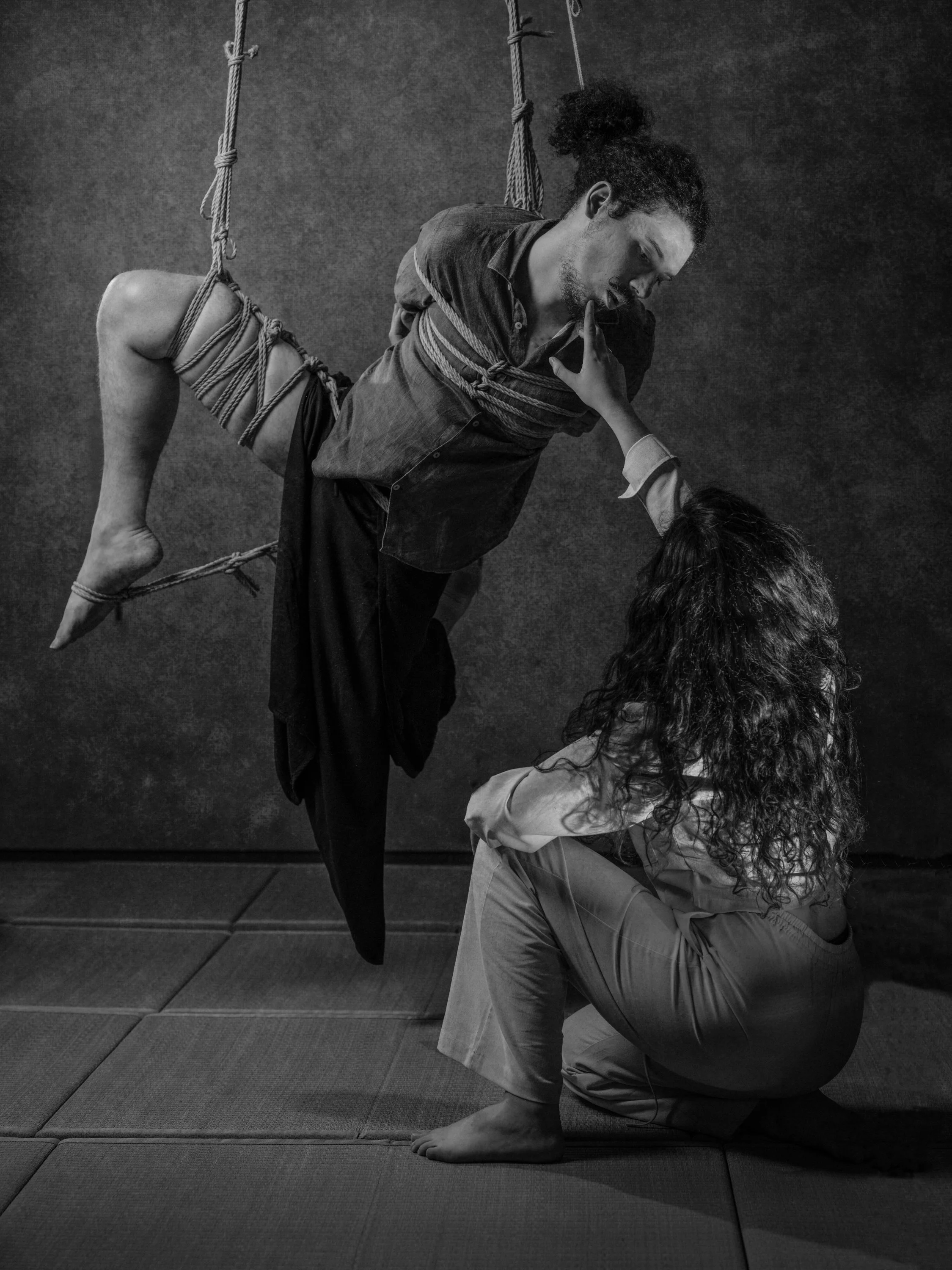 A person with curly hair kneels on the floor, holding another person upside down with ropes, in a studio with a plain background.