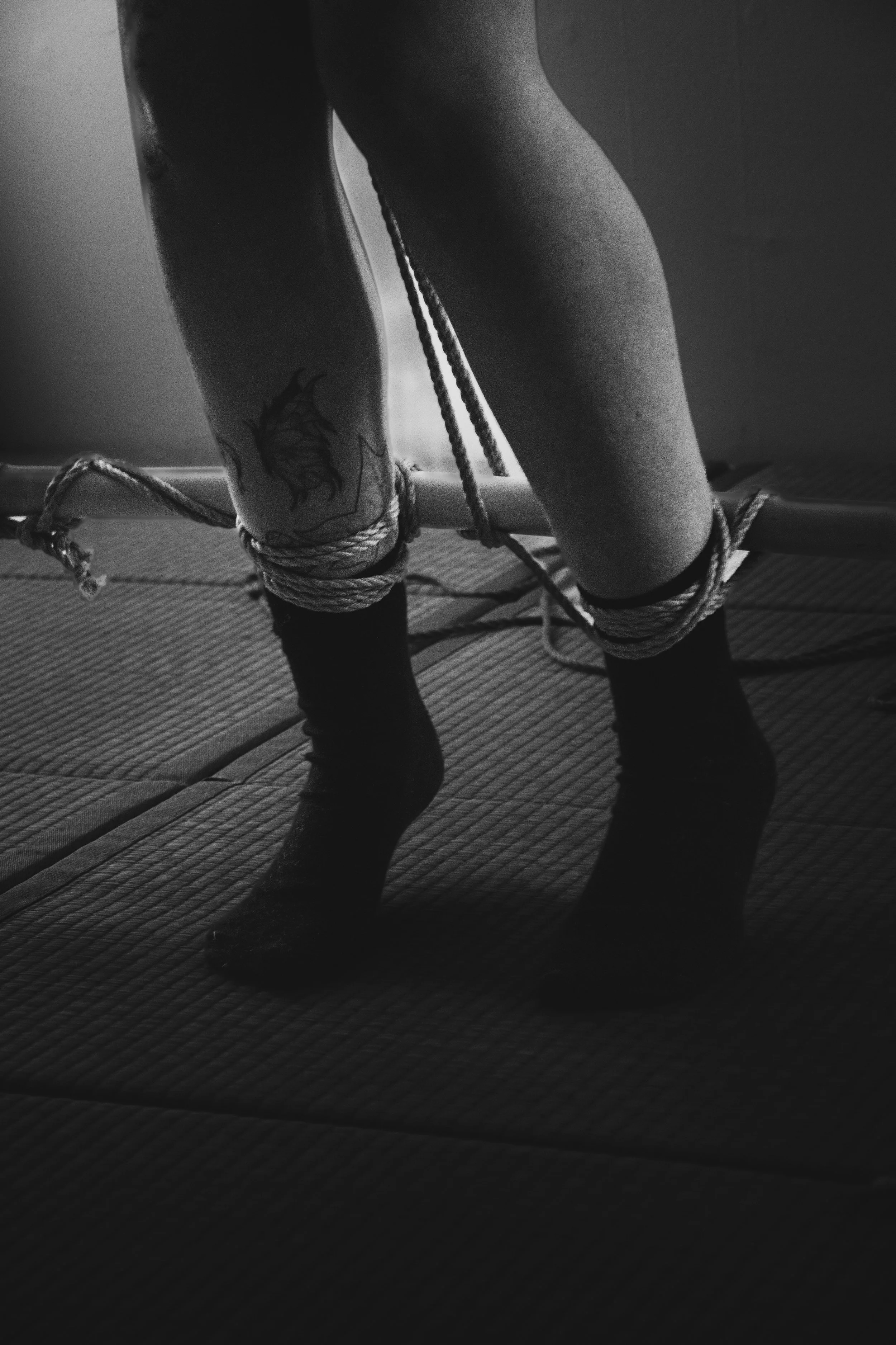 A person with tattoos is tying their ankles with rope while standing on a textured mat, with the ropes wrapped around their legs and possibly securing them to a bar or similar support.