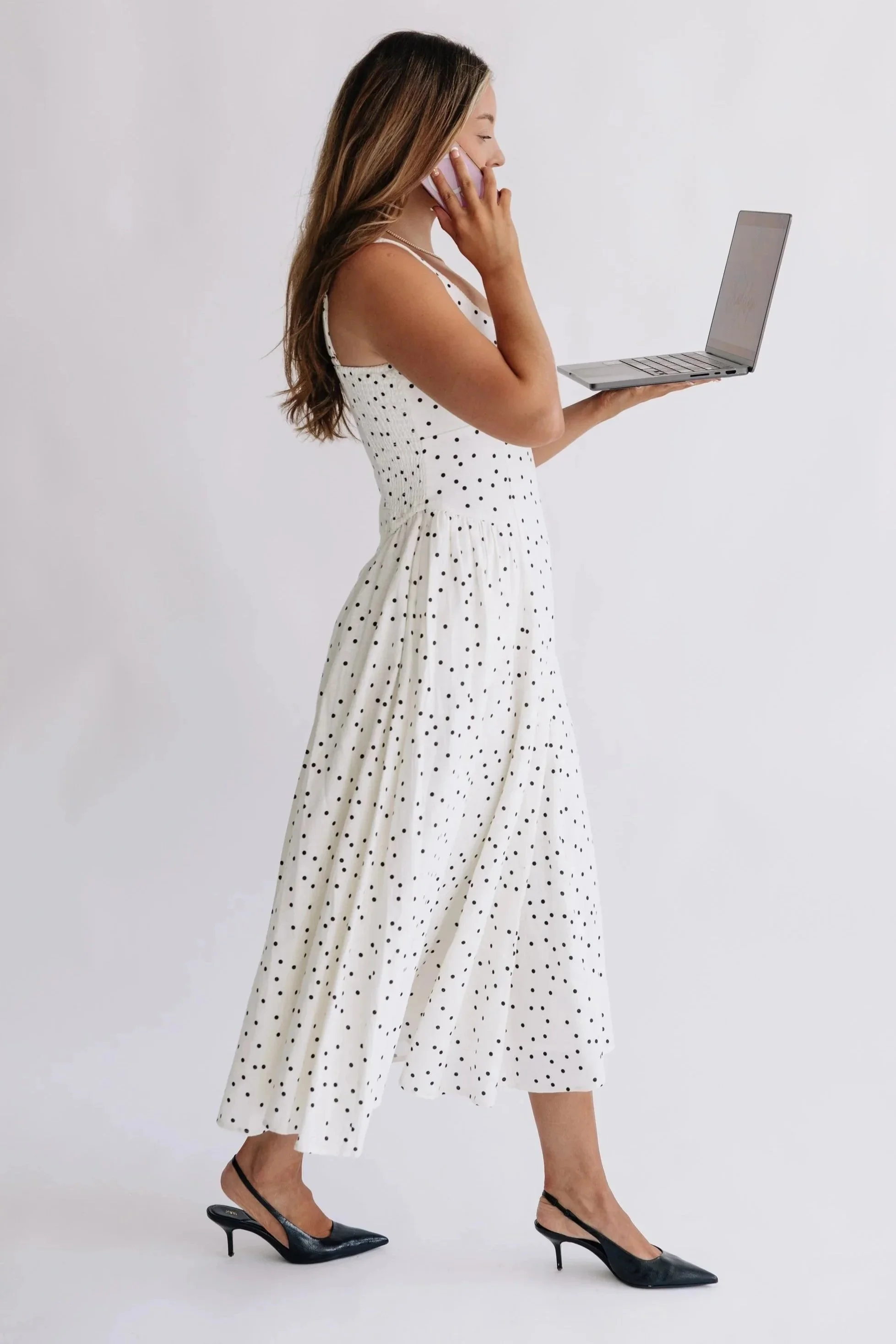 A woman in a white, sleeveless, polka dot dress with a flowing skirt, black high heels, holding a silver laptop in one hand, and talking on a pink phone. She is standing against a plain white background.