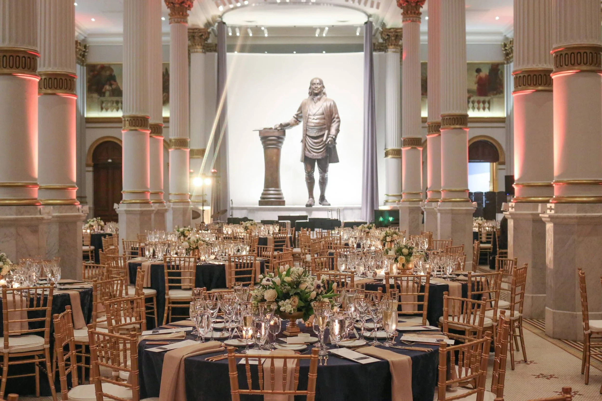 Elegant banquet hall with round tables set with glasses, plates, and floral centerpieces. The hall features tall, white columns with gold accents and a large statue of Benjamin Franklin in the background.
