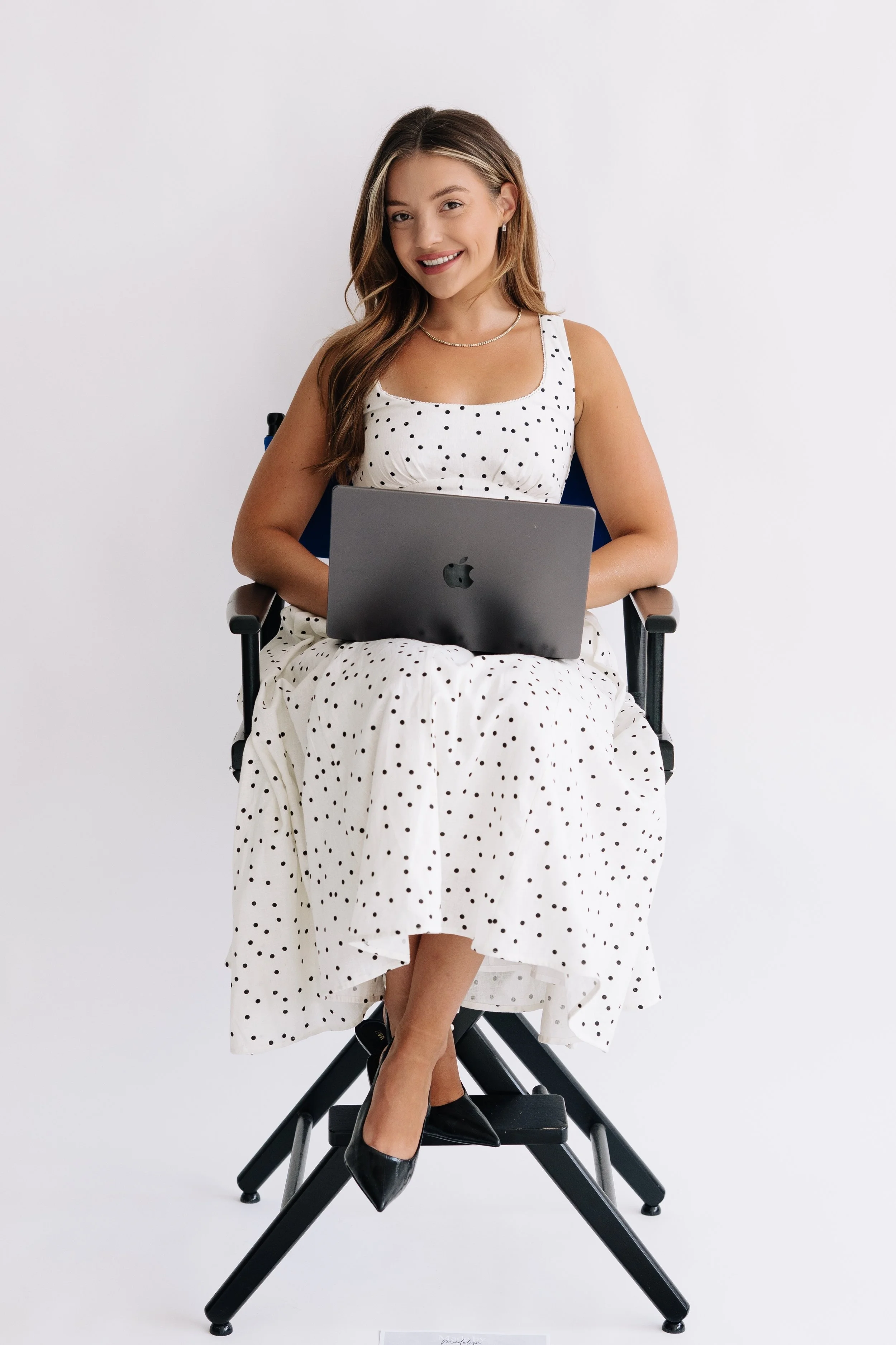 A woman sitting on a tall black chair with crossed legs, holding a silver MacBook laptop on her lap, wearing a white dress with black polka dots and black high heel shoes, smiling, against a plain white background.