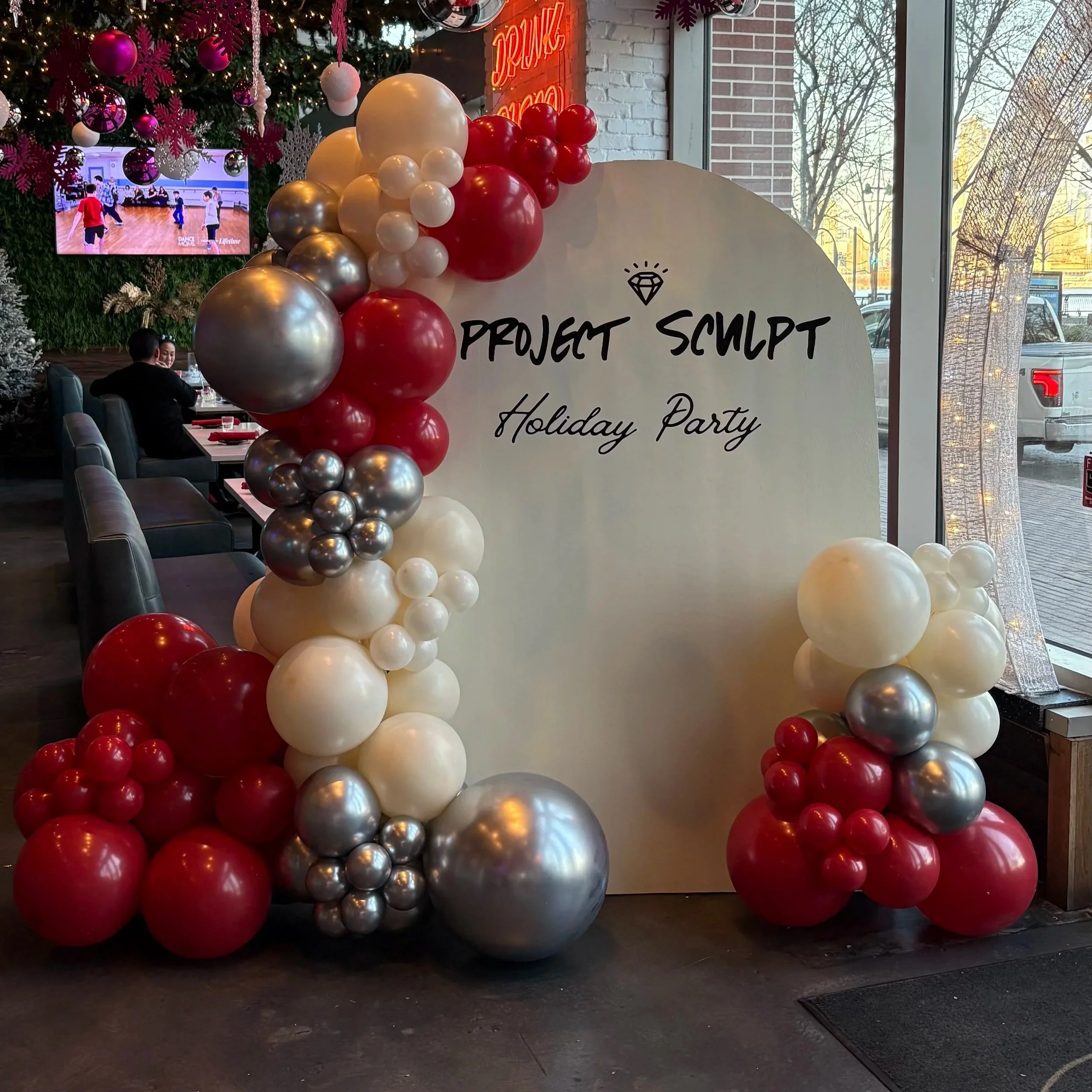 Decorative holiday balloon arrangement with red, white, silver, and pink balloons, a Christmas tree, wall-mounted TV displaying a dance class, and a sign that reads 'Project Sculpt Holiday Party' inside a festive venue with large windows showing trees and parking outside.