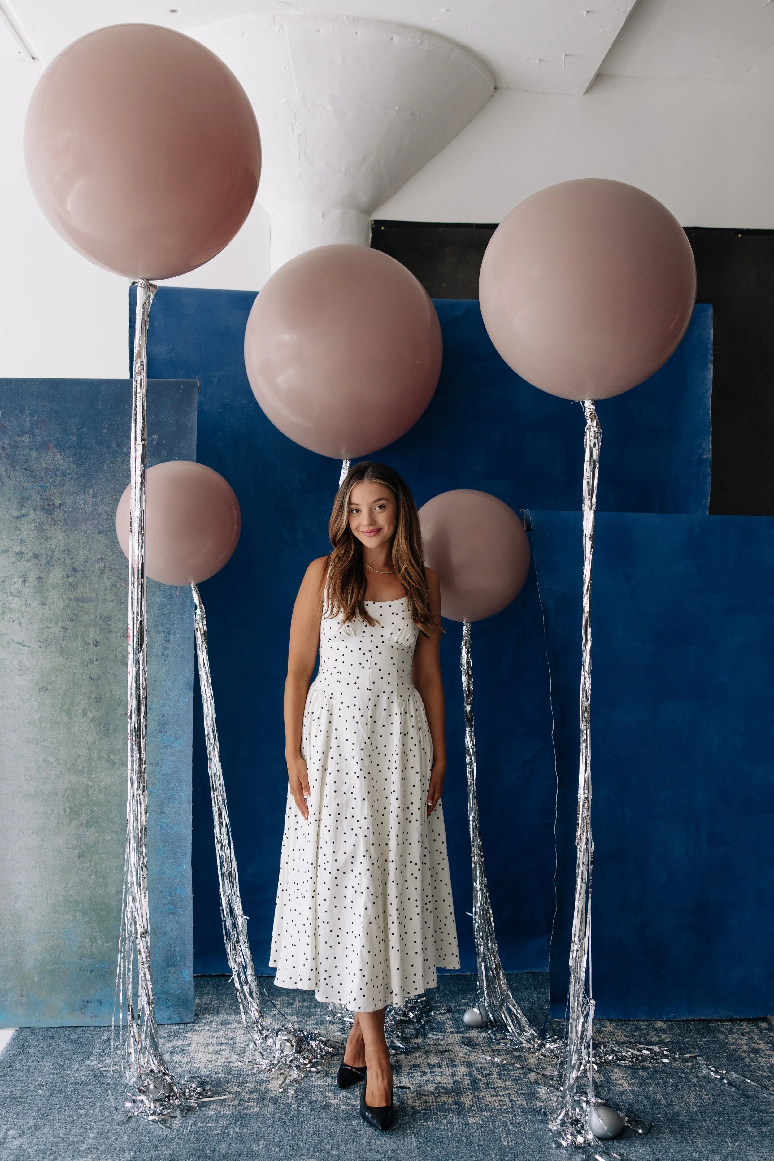 A woman in a white polka dot dress standing among large pink balloons with silver ribbon strings in a studio with a blue and white background.