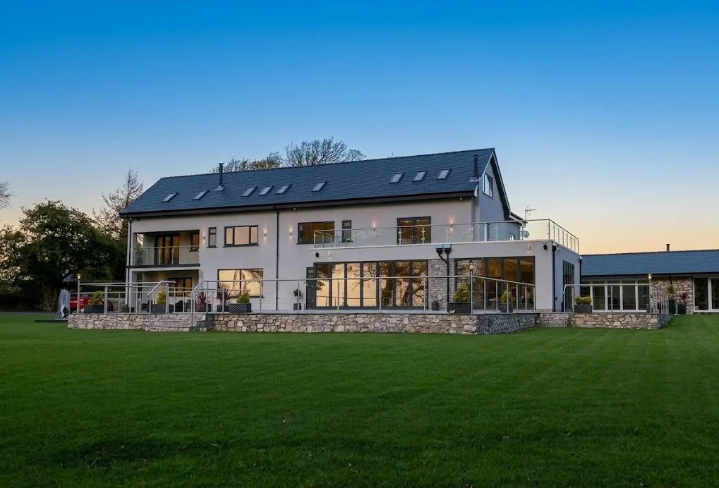 Modern two-story house with large windows, a balcony, and a spacious green lawn at sunset.