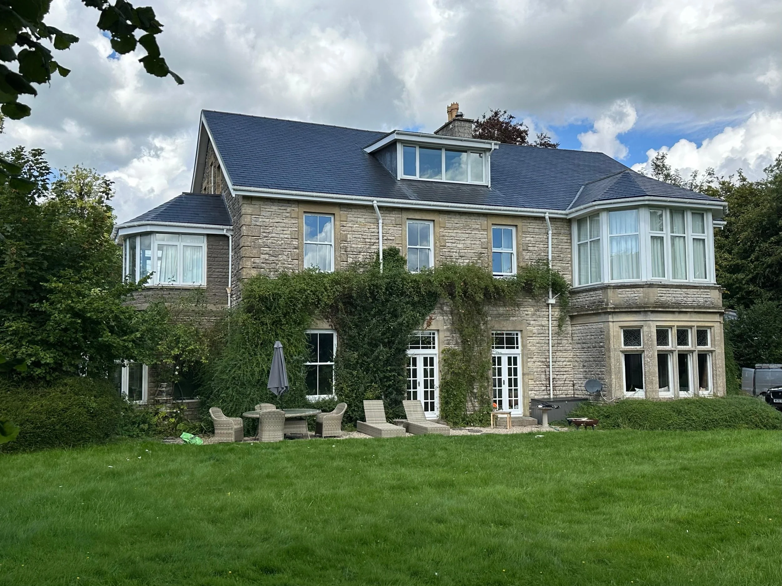 A large stone house with a dark blue roof, multiple windows, a patio with patio furniture, and a green lawn, under a partly cloudy sky.