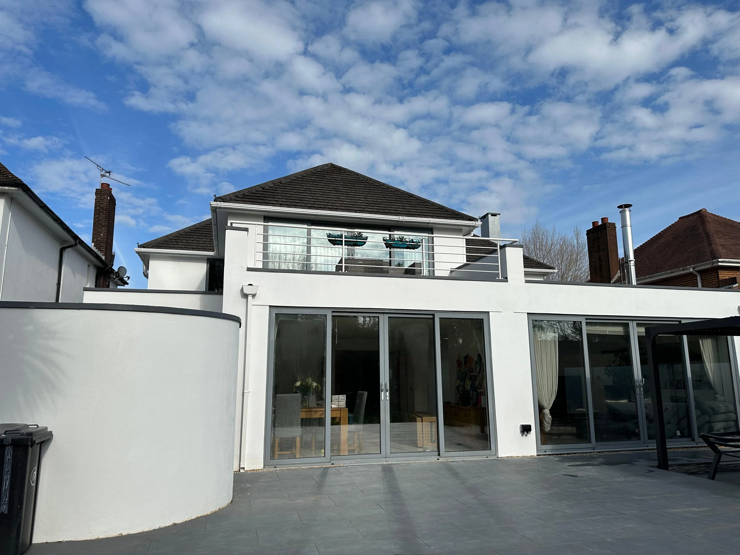 Rear view of a modern house with large glass doors, a balcony with potted plants, and a clear blue sky with clouds.