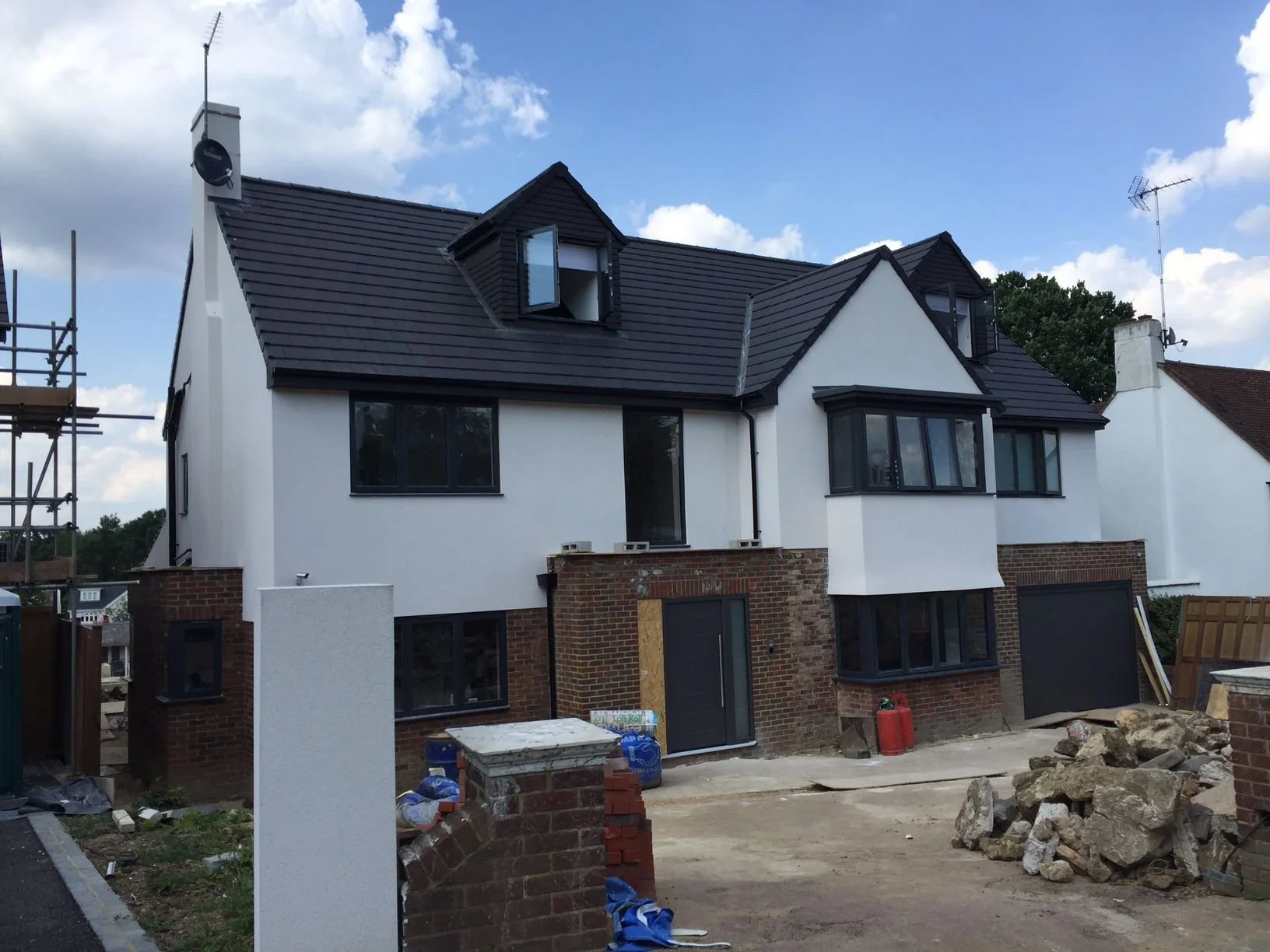 Under construction residential house with black roof, white walls, and brick foundation, surrounded by construction materials and debris.