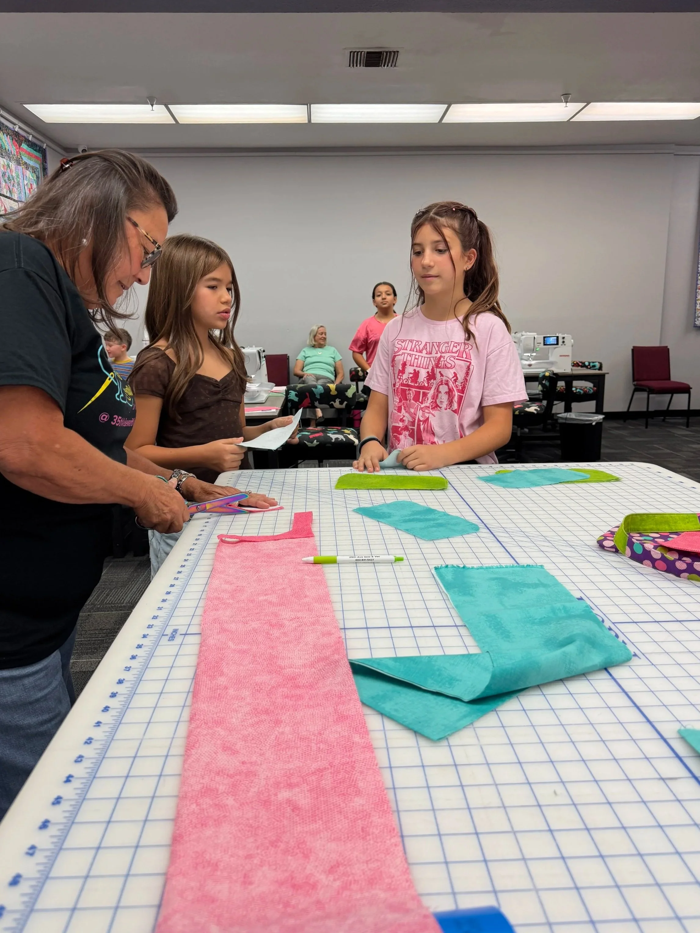 Children and an adult working together on a sewing project at a table with fabric pieces and tools, in a room with chairs and sewing machines in the background.
