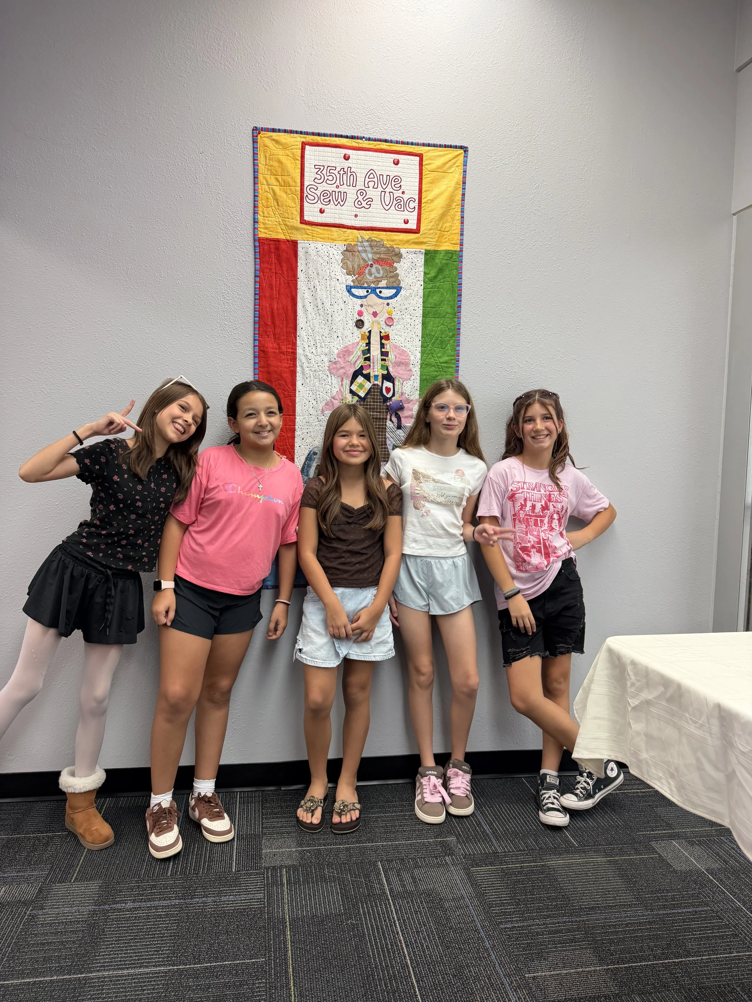 Five young girls posing in front of a colorful quilt with the text "35th Ave Sew & Vac".