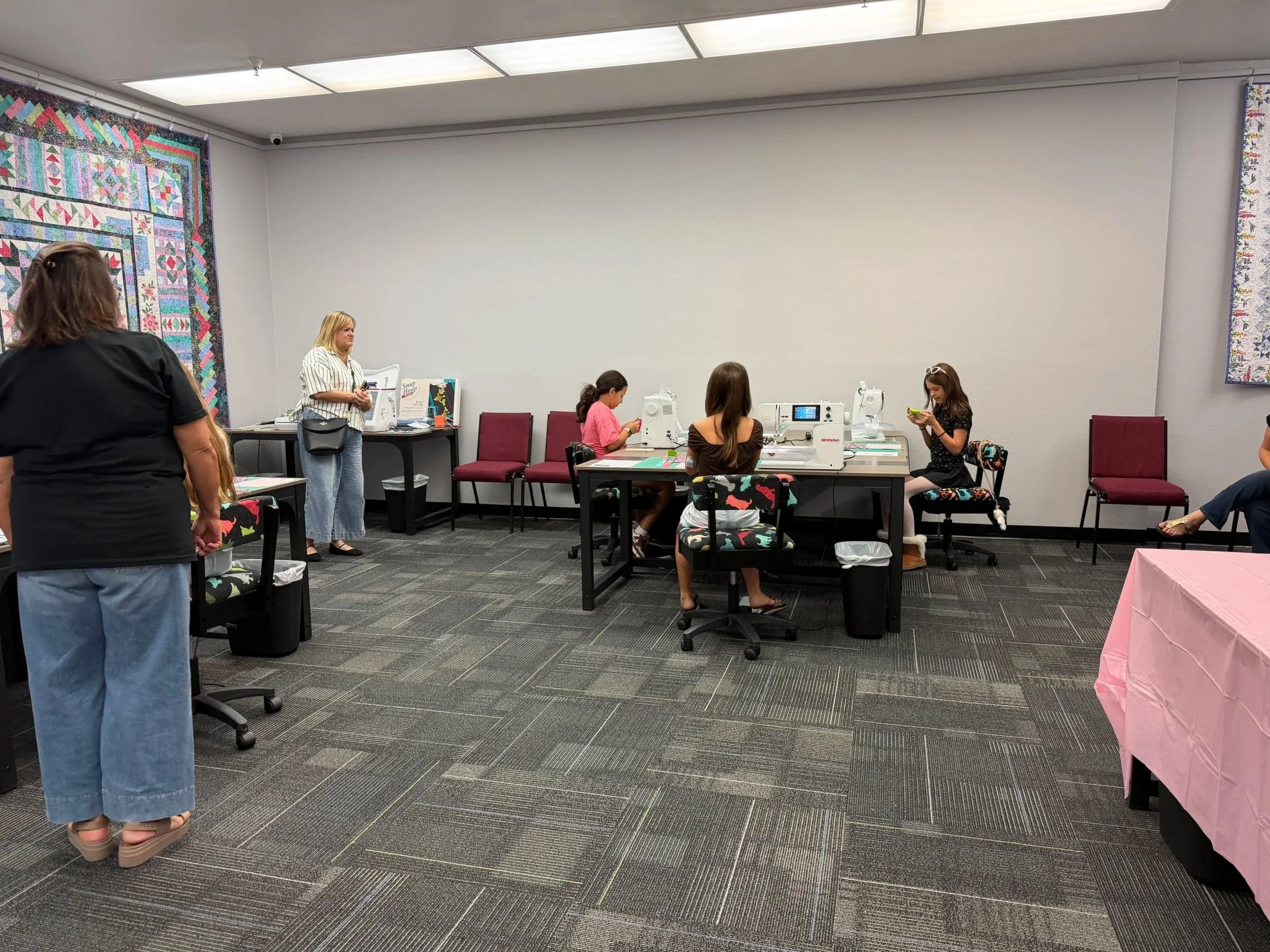A quilting workshop with several people, including children, gathered around sewing machines in a room decorated with colorful quilts on the walls.