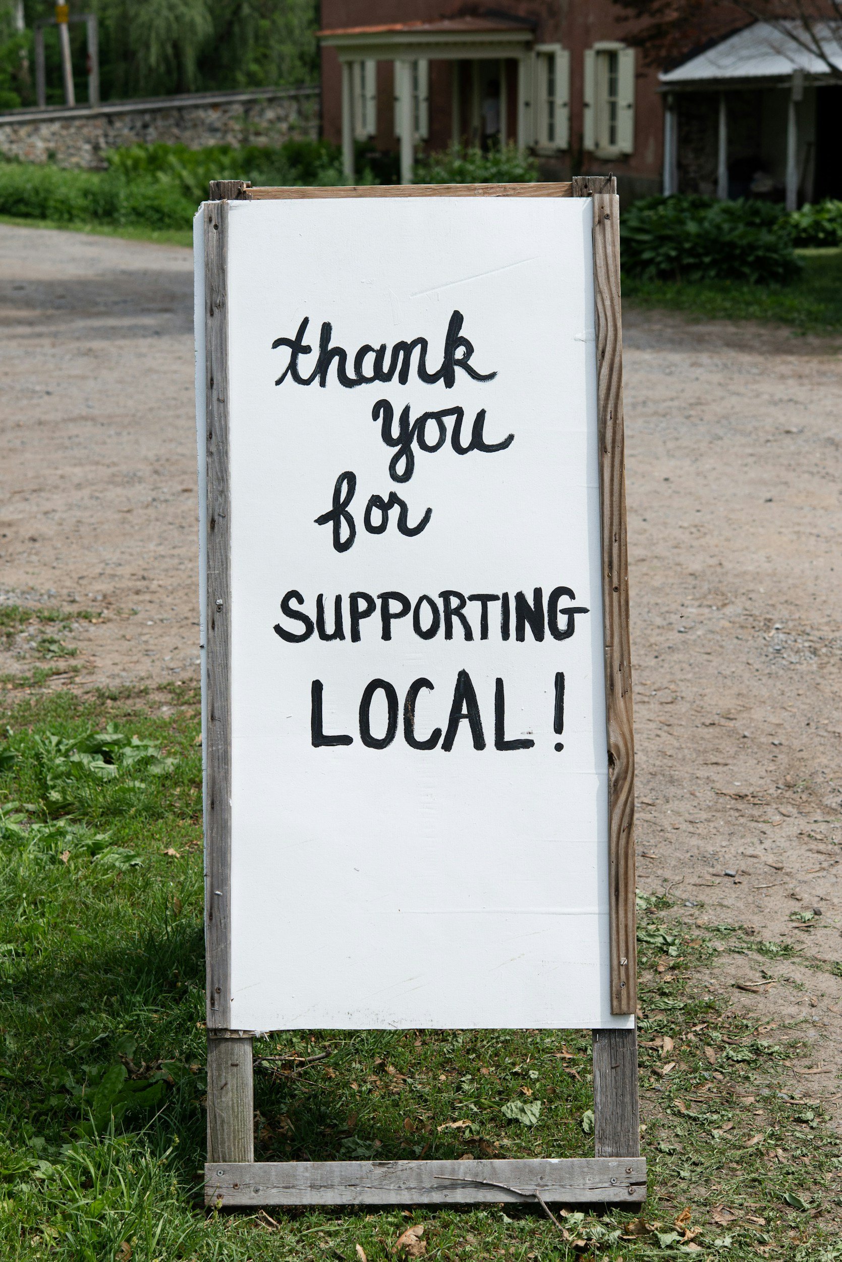 White signboard with a wooden frame displaying the message, 'Thank you for supporting local!' in black handwritten font, situated on a grassy area near a dirt pathway, with houses and greenery in the background.