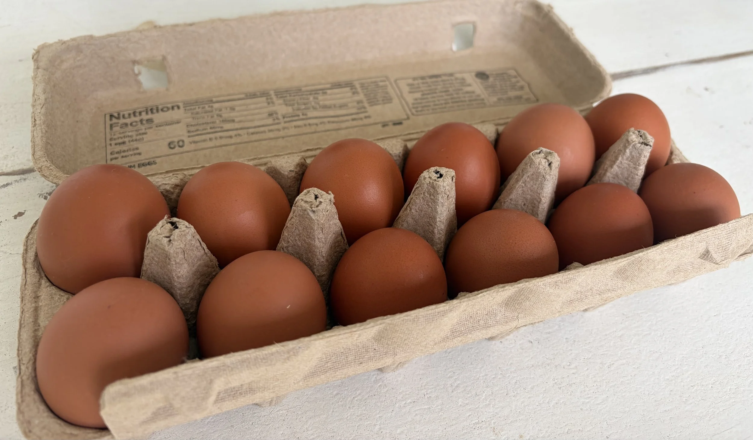 A cardboard egg carton with 12 brown eggs inside, placed on a white surface, with a partial view of the carton lid showing nutrition facts.