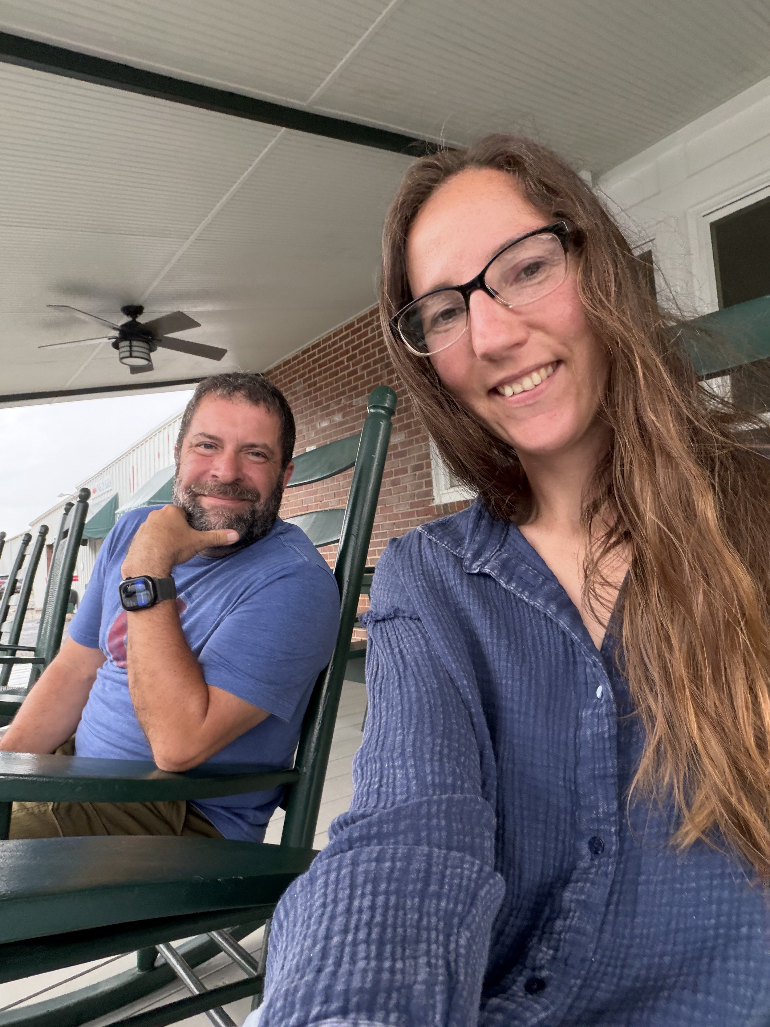 A woman with long brown hair and glasses taking a selfie with a man with a short beard and a blue shirt sitting outside at a table with green chairs on a porch with a brick wall and ceiling fan in the background.