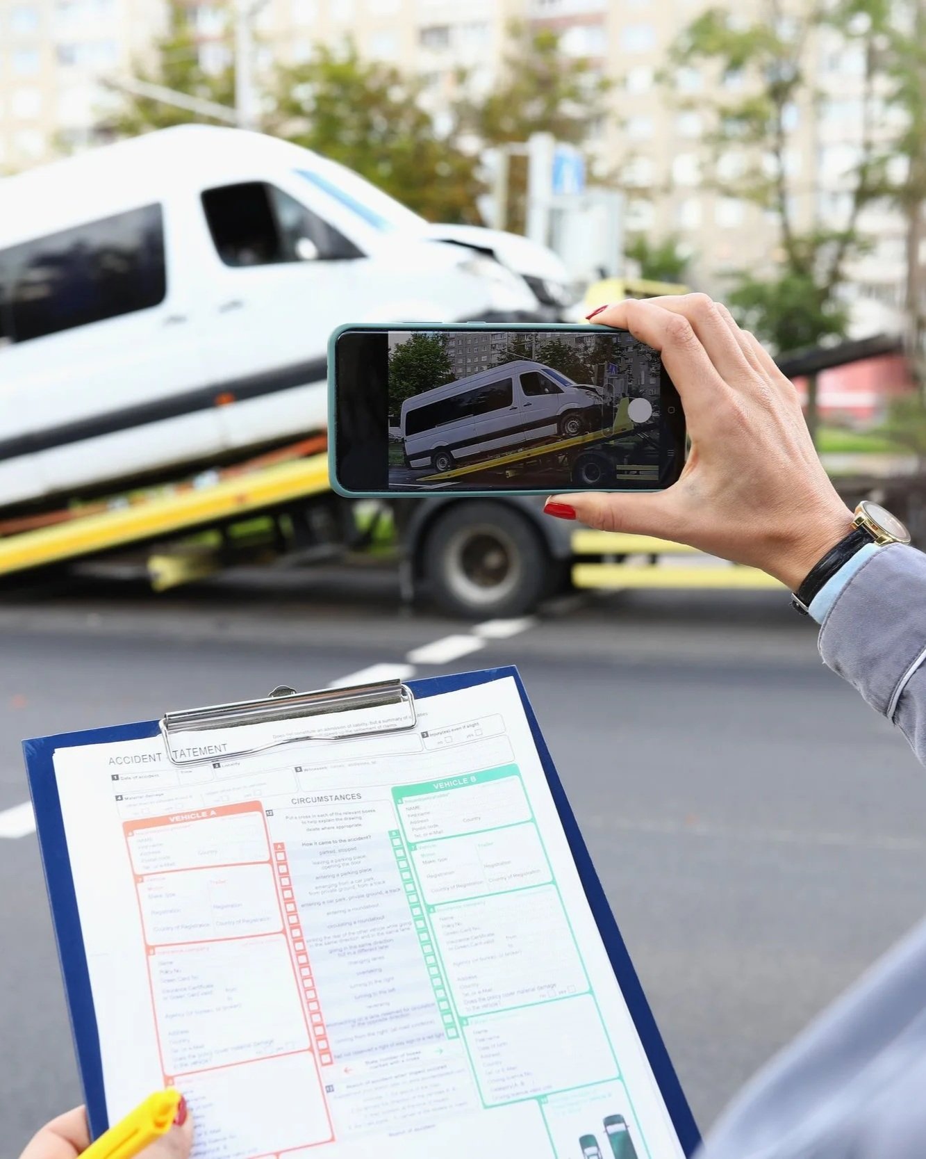 A person holding a yellow pen and an accident statement form in front of a tow truck carrying a white van.