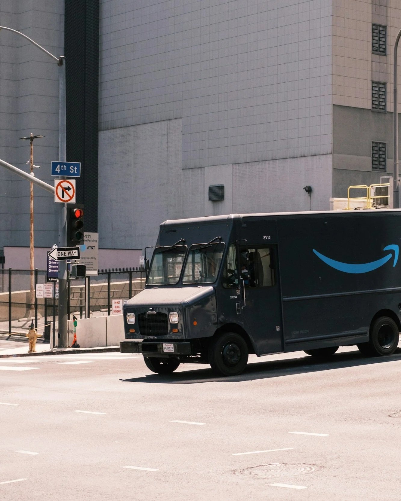 A black Amazon delivery truck driving on a city street, with a large gray building, traffic lights, and street signs in the background.