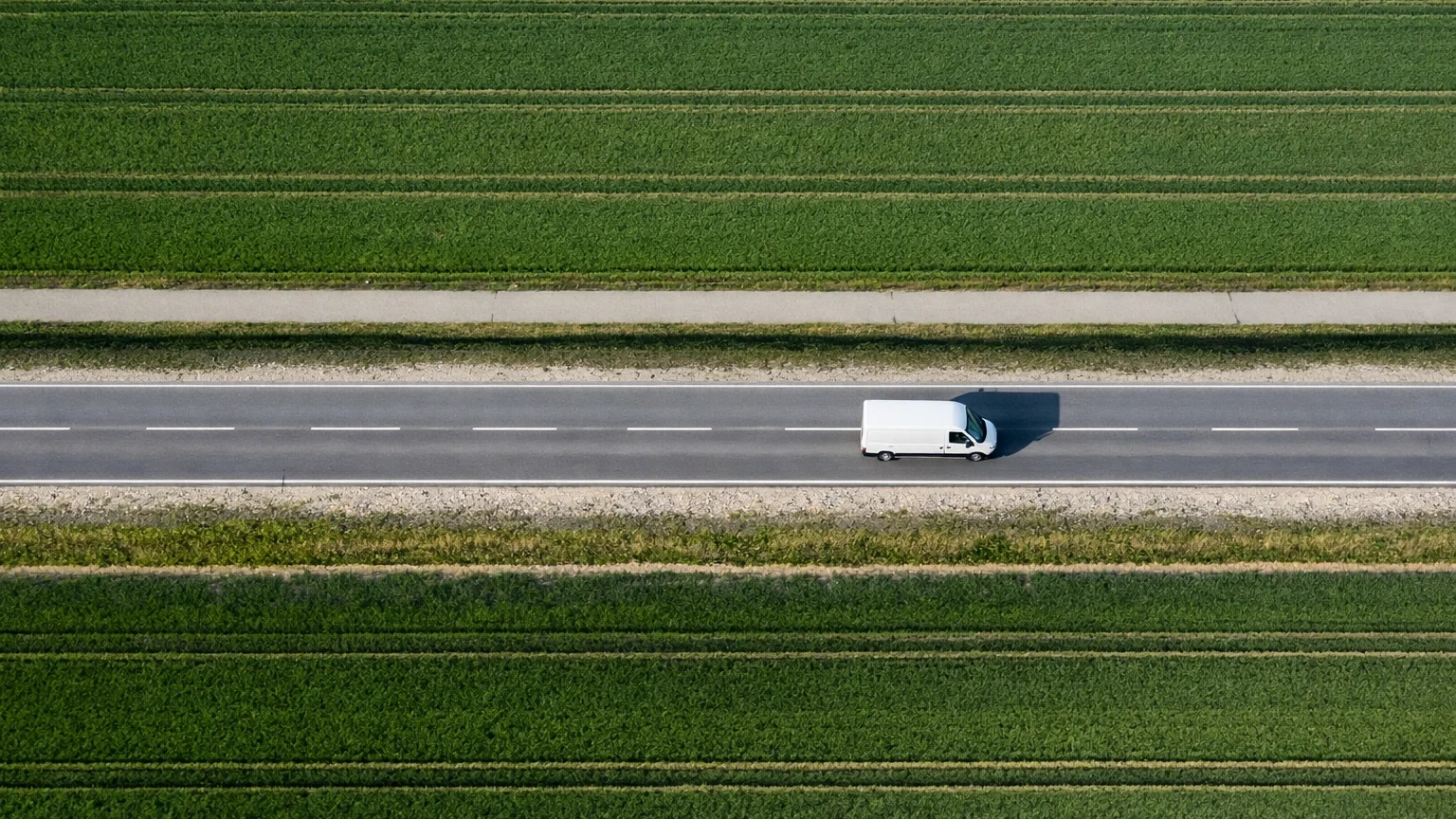 An aerial view of a white van driving on a two-lane asphalt road, with green fields on either side.