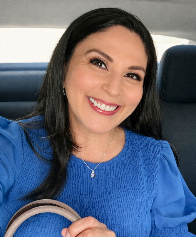 A woman with dark hair smiling while sitting in a car, wearing a blue top and a silver necklace.