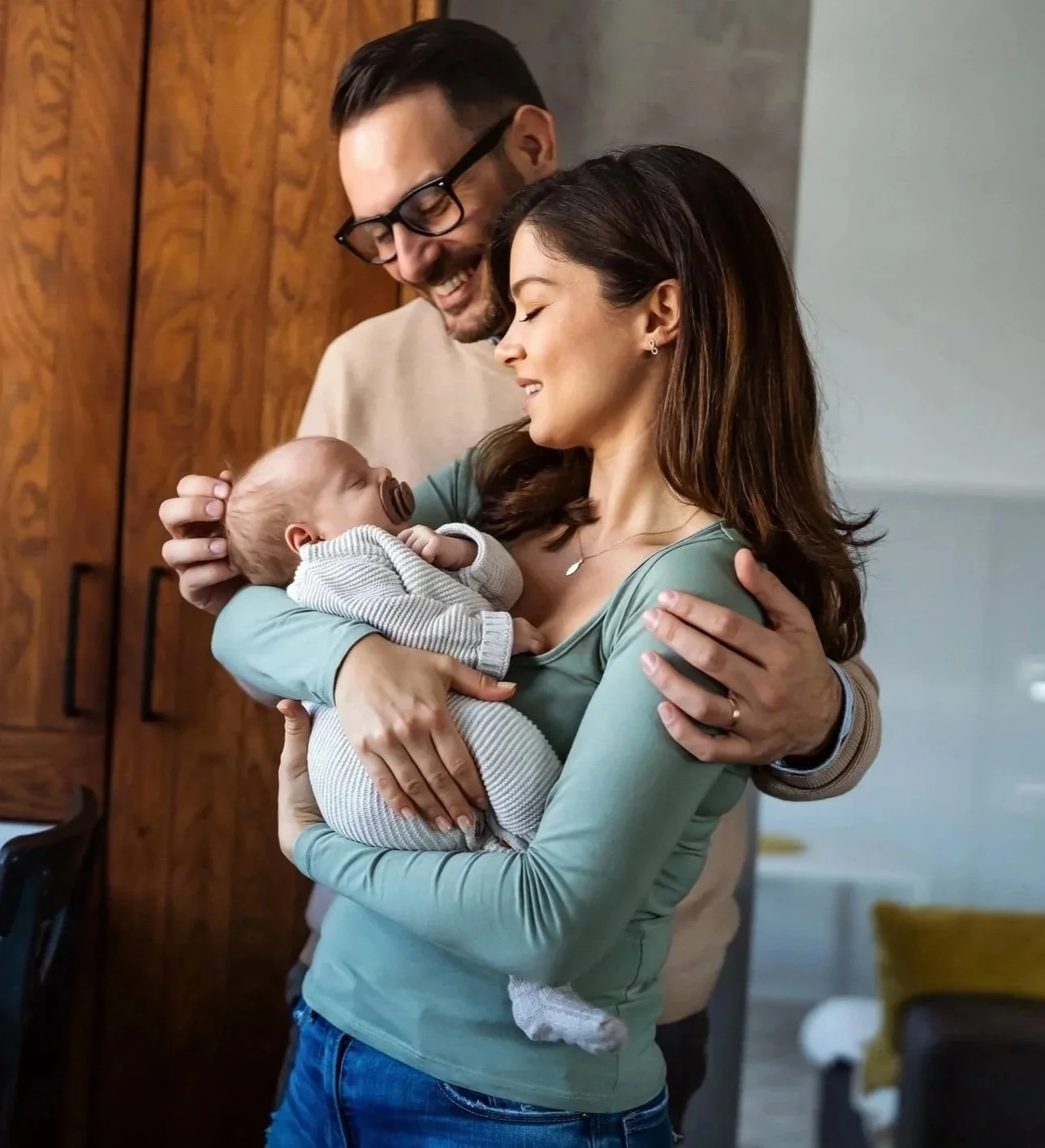 A couple standing together indoors, gently holding and looking down at their sleeping newborn baby, who is wrapped in a light gray knit outfit with a pacifier.