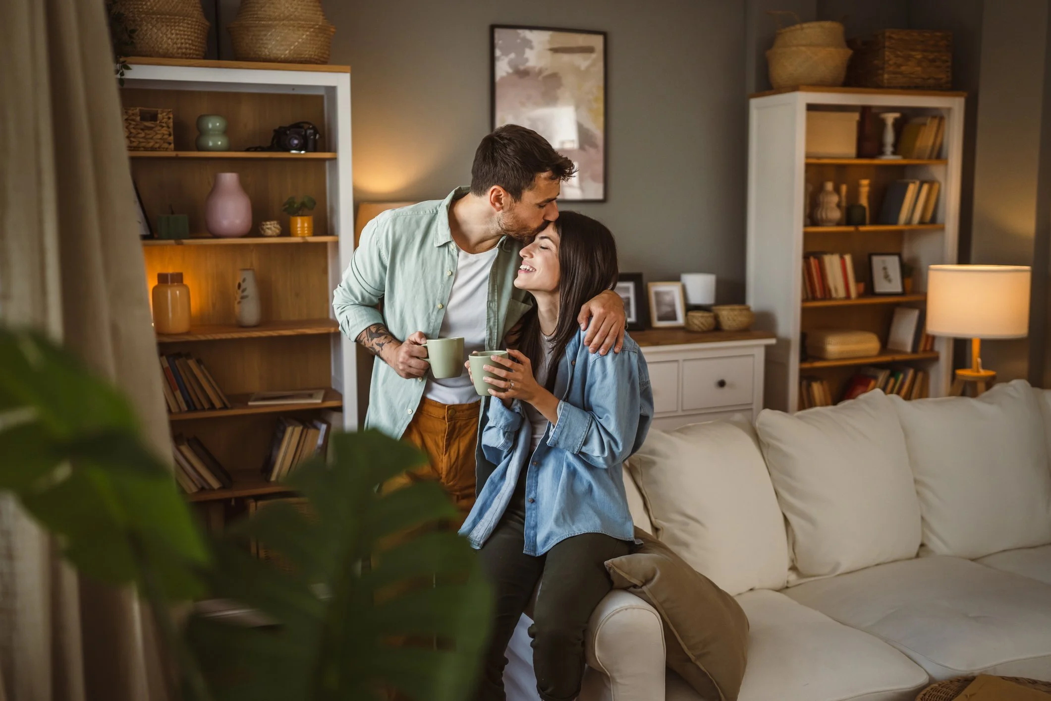 A couple sharing a tender moment in a living room, holding coffee mugs and smiling lovingly at each other.