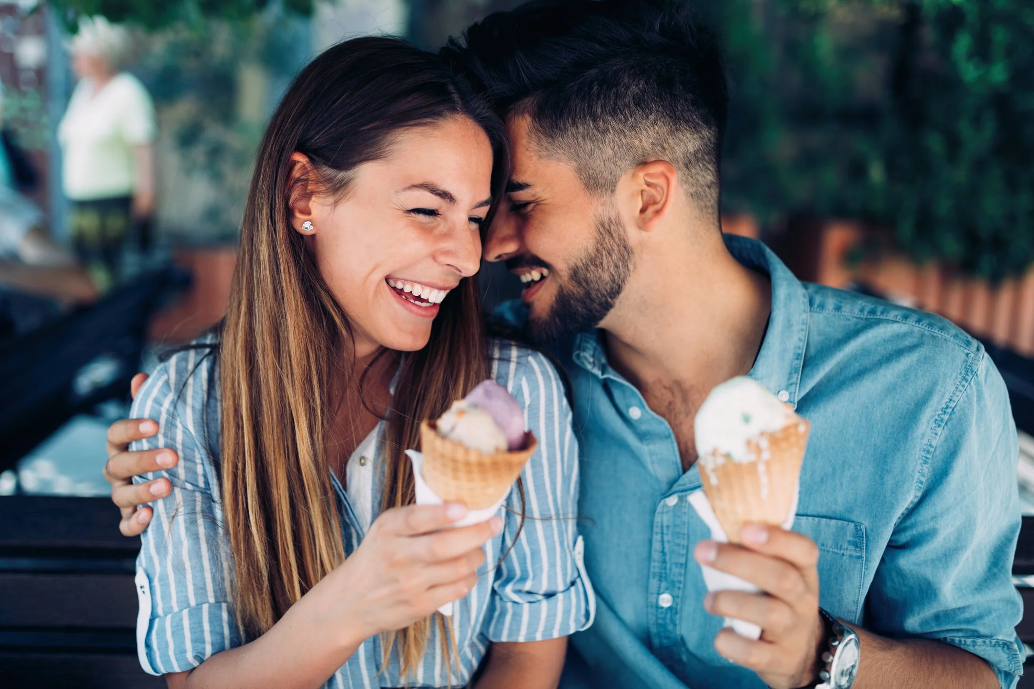 A smiling young couple sitting close together outdoors, each holding an ice cream cone, laughing and leaning their heads together affectionately.