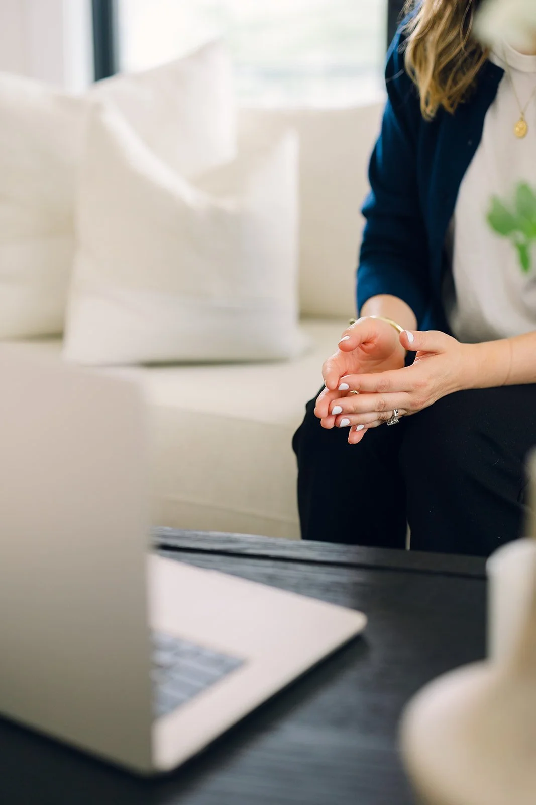 Close-up of a woman's hands with manicured nails, wearing a ring, sitting at a table with a laptop, in a bright room.