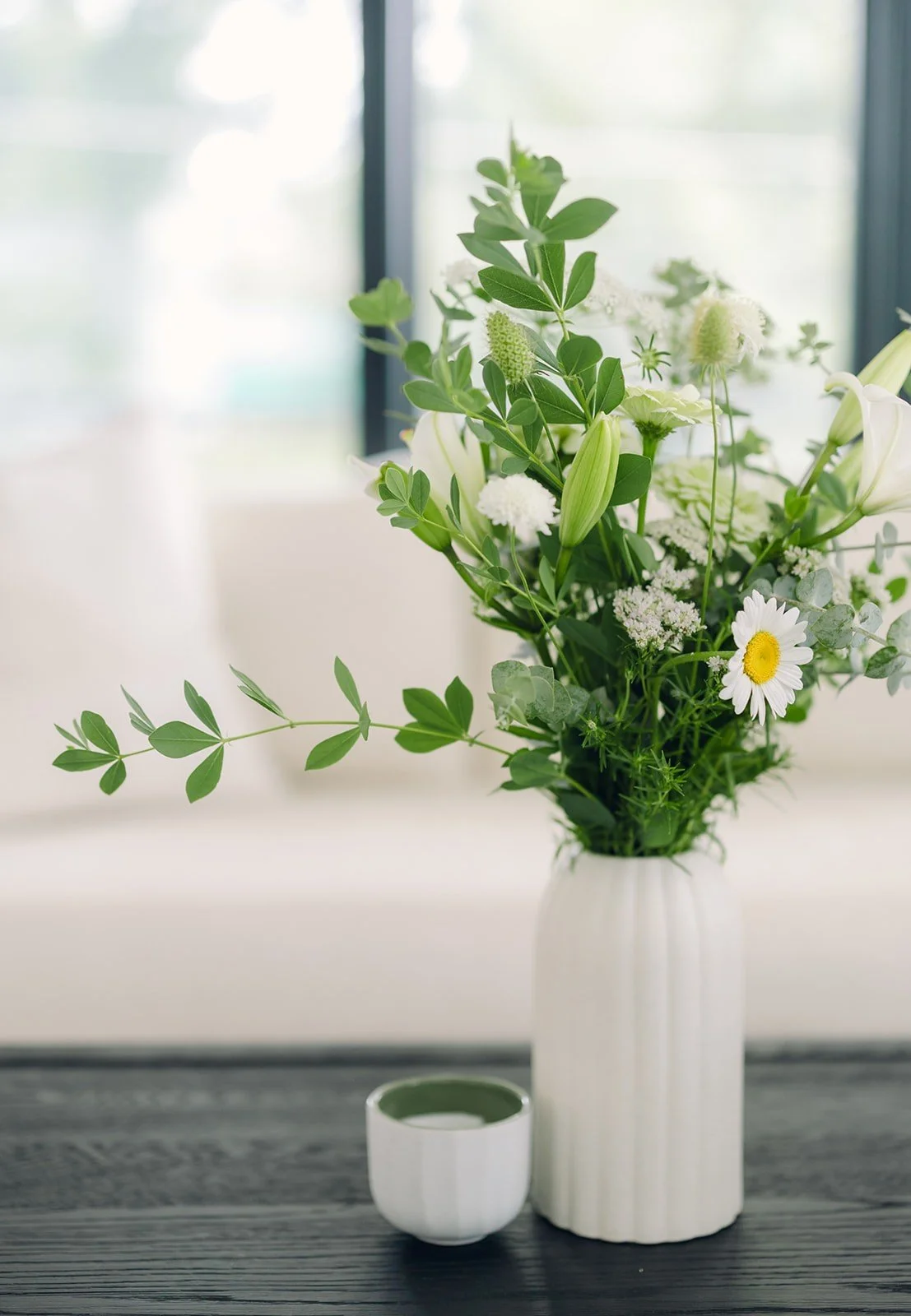 A white vase filled with green leafy flowers and white daisies on a dark wooden table near a window.