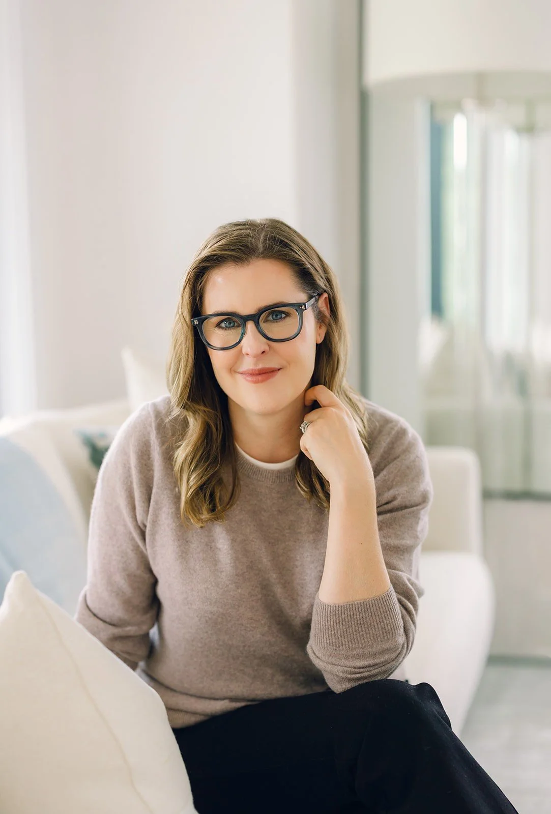 Trauma therapist Claire Connolly, LICSW is wearing glasses, a beige sweater, and black pants, sitting on a light-colored sofa in a bright room, smiling at the camera.
